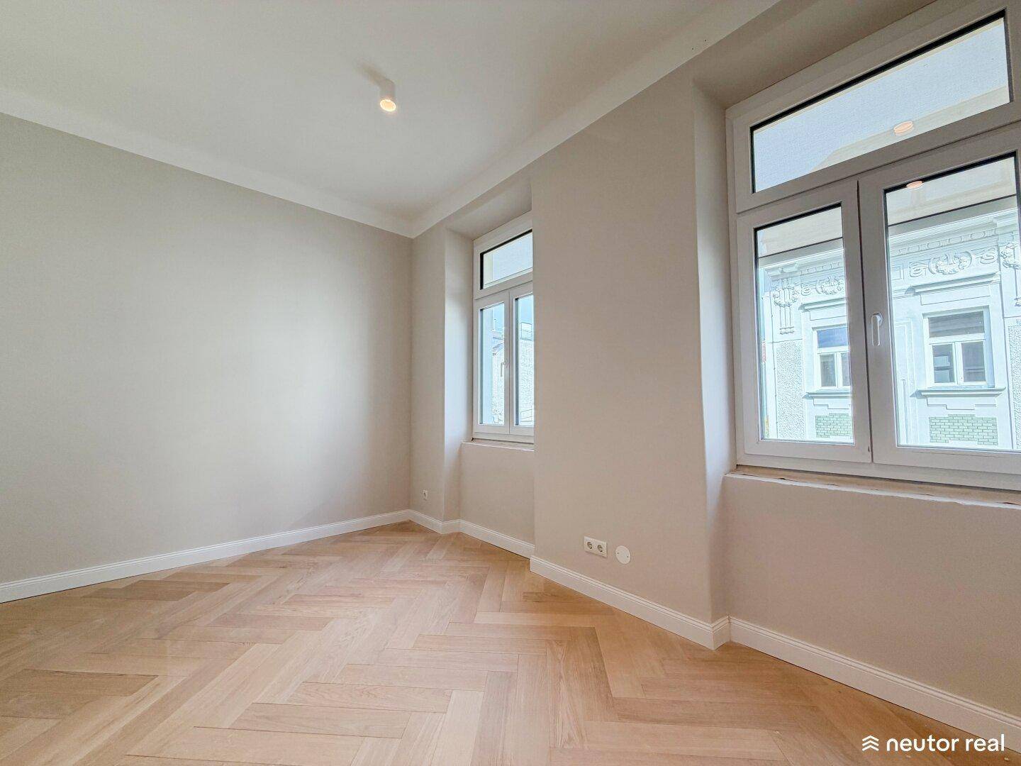 Geräumiges Zimmer mit Fischgrätparkett und großen Fenstern mit Blick auf die Stadtarchitektur.