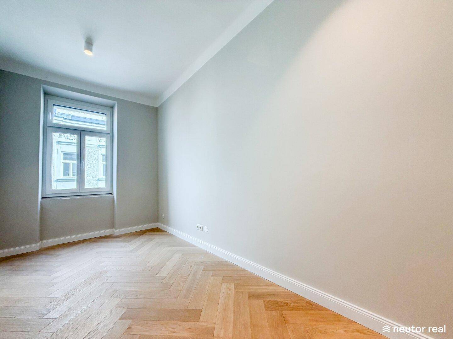 Helles Zimmer mit Fischgrätparkett und einem Fenster, das viel Tageslicht hereinlässt.