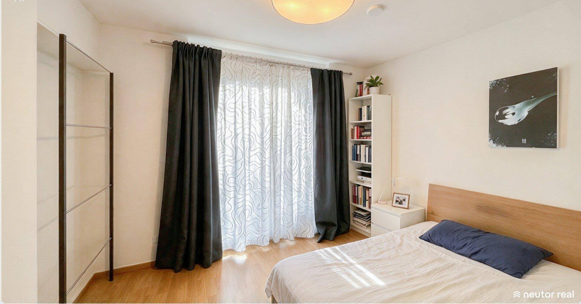 Helles Schlafzimmer mit Bett, Nachttisch und großem Fenster mit Vorhängen für viel Tageslicht.