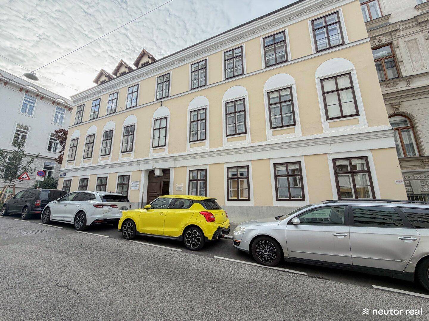 Historische Fassade eines Mehrfamilienhauses mit vielen Fenstern und parkenden Autos auf der Straße.