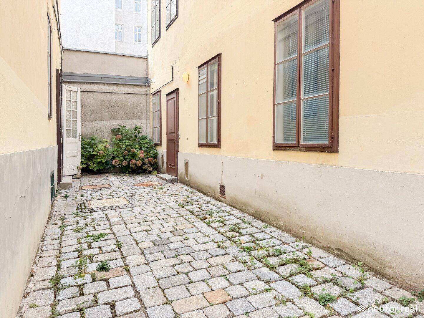 Schmaler Innenhof mit Kopfsteinpflaster und Pflanzen, der zu einem weiteren Bereich führt.