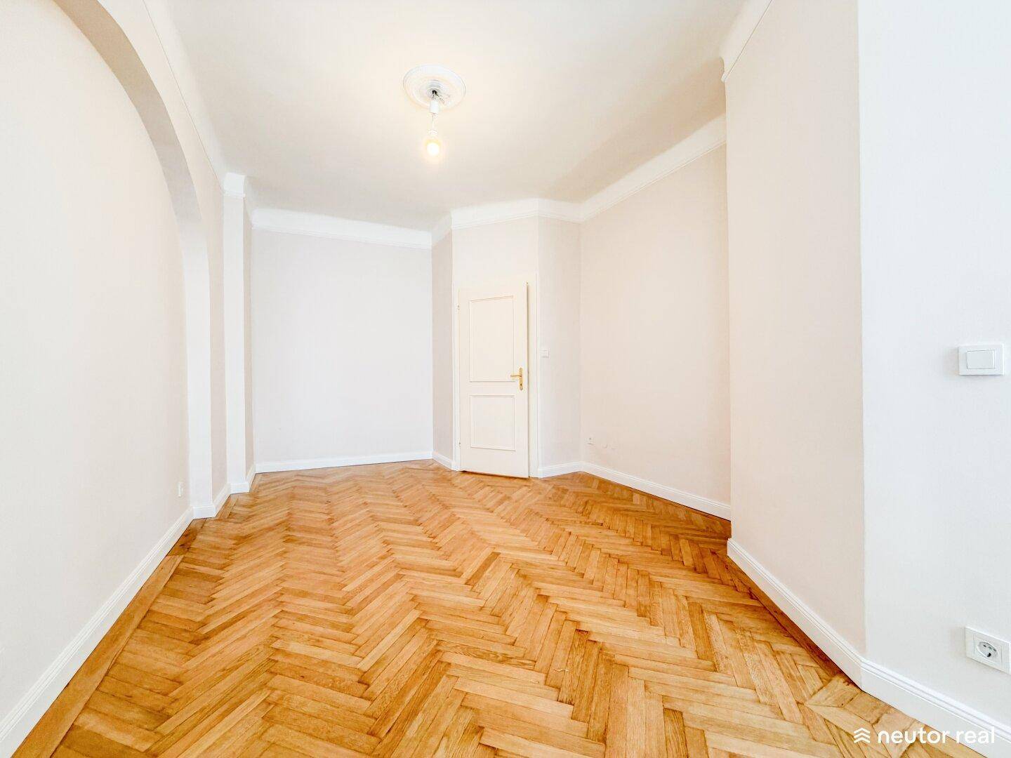 Helles Zimmer mit Fischgrätparkett, hohen Decken und einer Tür, die zu einem weiteren Raum führt.