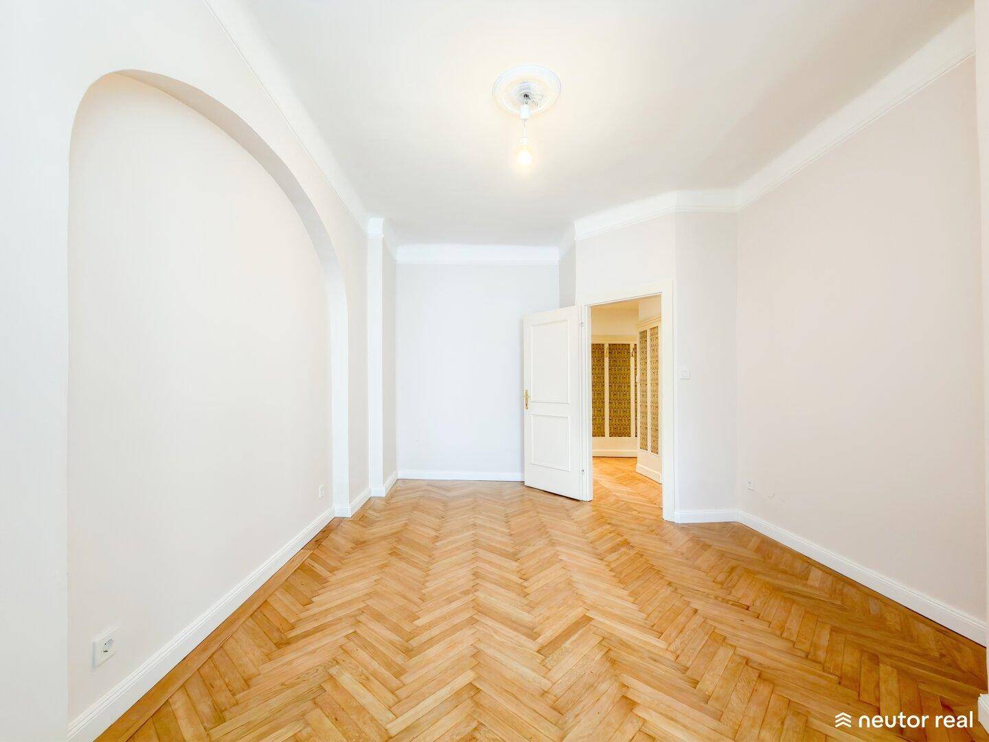 Leeres Zimmer mit Fischgrätparkett, hohen Decken und einem eleganten Bogen als Durchgang.