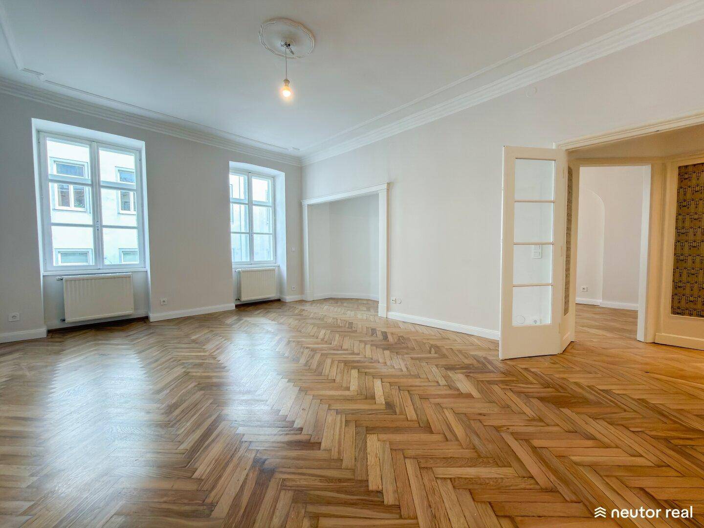 Großes Wohnzimmer mit Fischgrätparkett, hohen Decken und zwei hohen Fenstern für viel Licht.