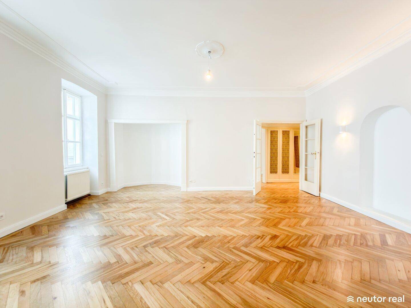 Geräumiges Wohnzimmer mit Fischgrätparkett, hohen Decken und großen Fenstern für viel Tageslicht.