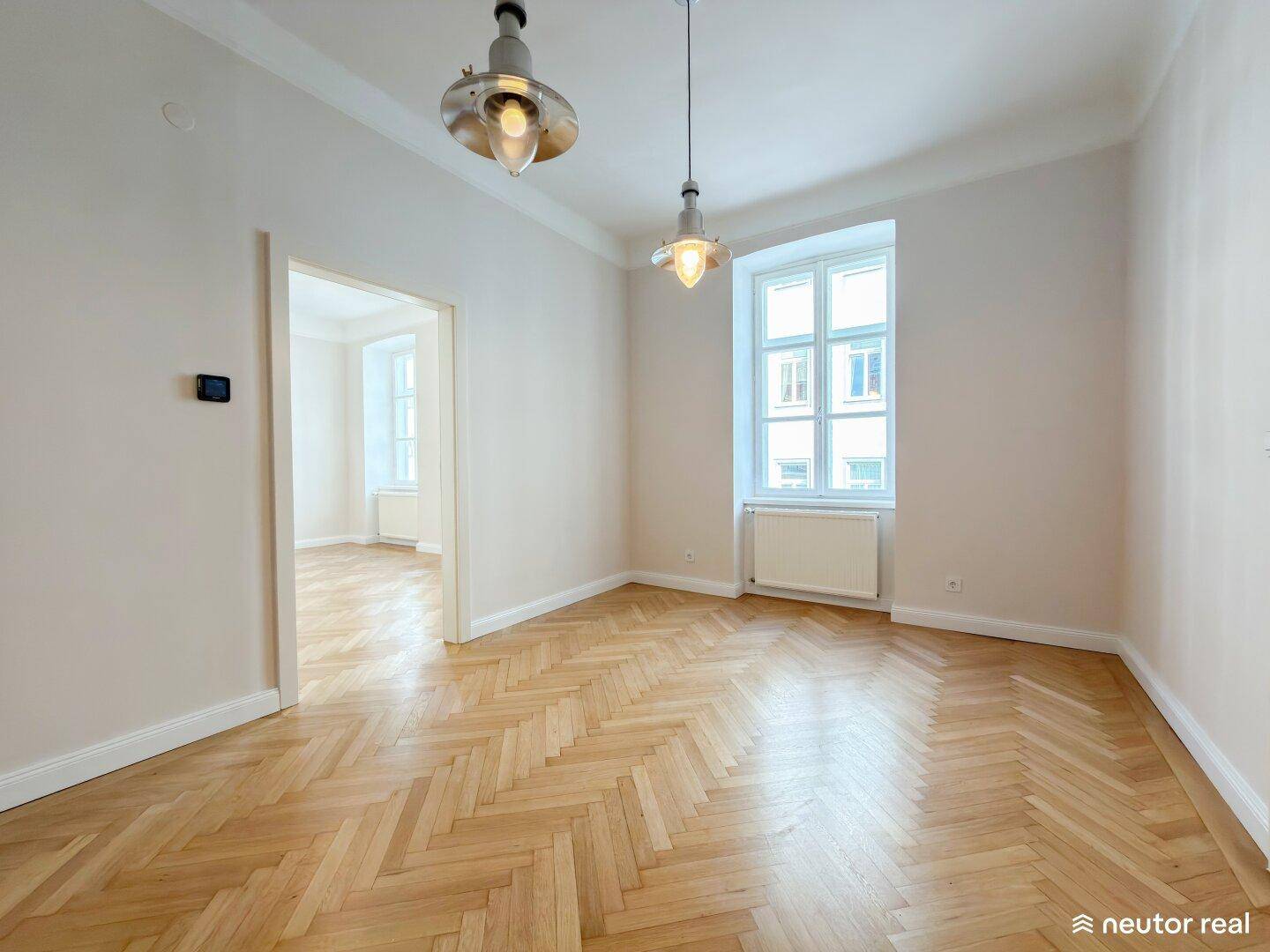 Helles Zimmer mit Fischgrätparkett, hohen Decken und einem großen Fenster, das viel Licht hereinlässt.