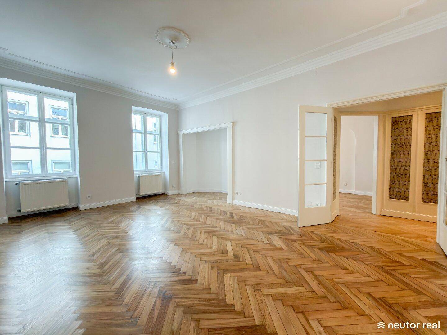 Helles Wohnzimmer mit Fischgrätparkett, hohen Decken und großen Fenstern, die den Raum erhellen.
