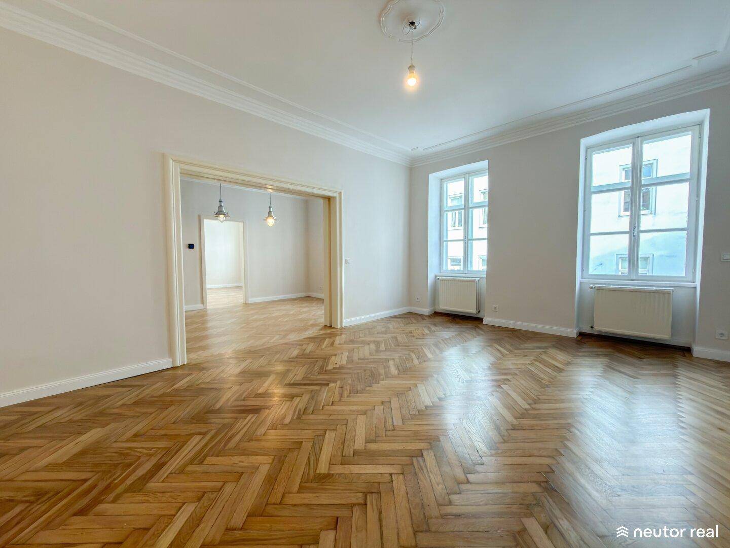 Geräumiges Wohnzimmer mit Fischgrätparkett, hohen Decken und zwei großen Fenstern.