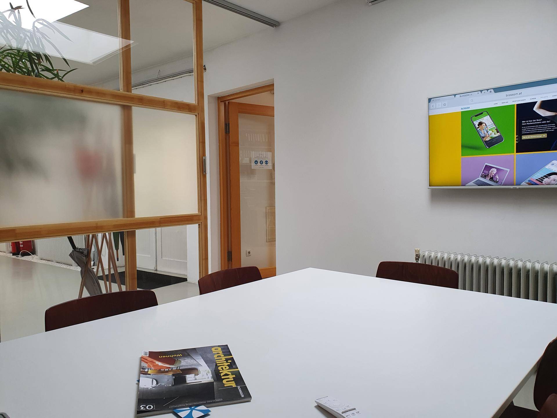 Moderner Konferenzraum mit großem weißen Tisch, Stühlen und einem an der Wand montierten Bildschirm für Präsentationen.