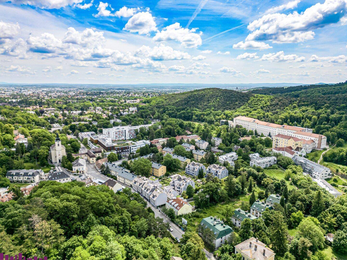 Weitläufige Luftaufnahme der Stadt und der umliegenden grünen Hügellandschaft unter blauem Himmel.