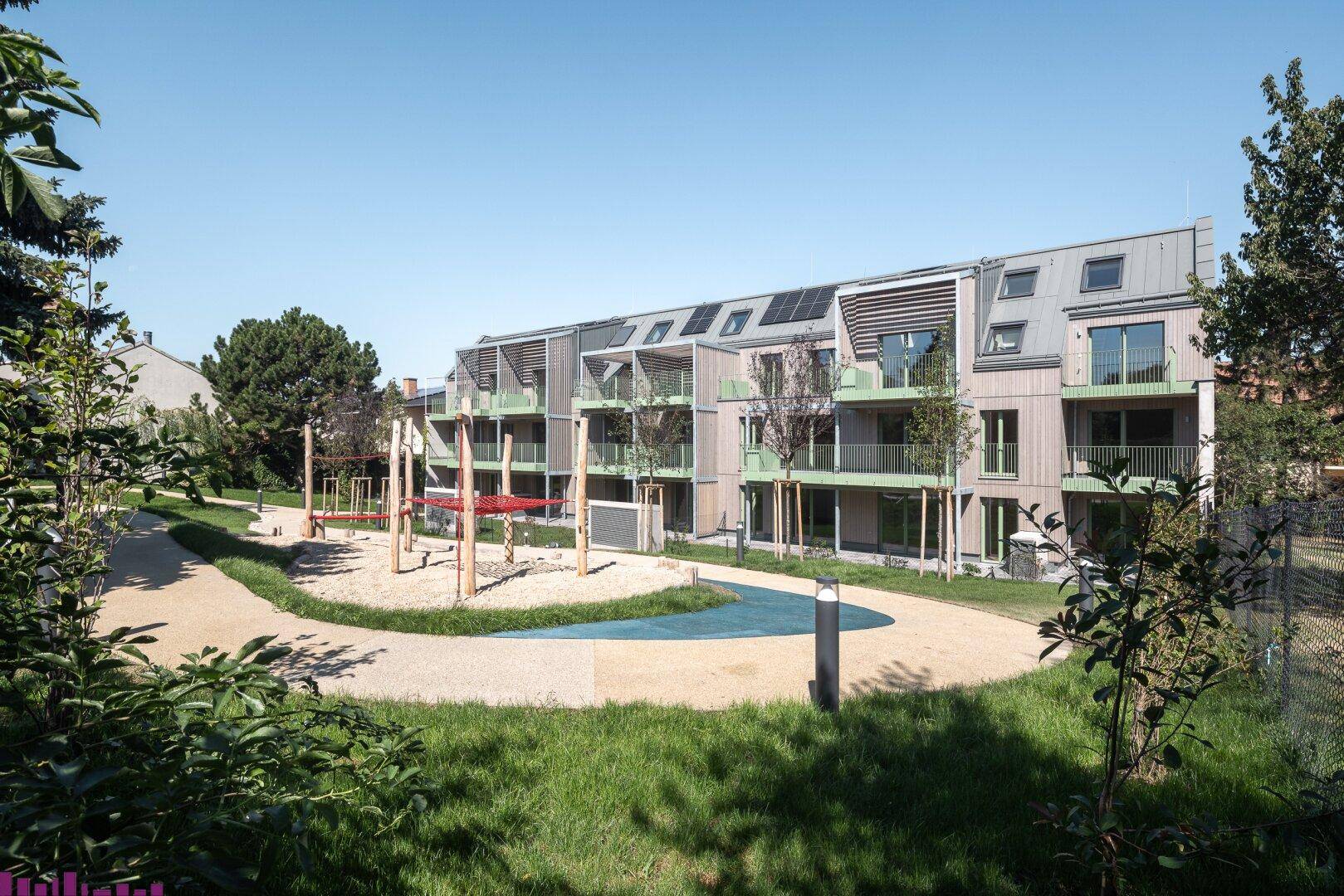 Moderne Wohnanlage mit Gemeinschaftsgarten und Kinderspielplatz unter blauem Himmel.