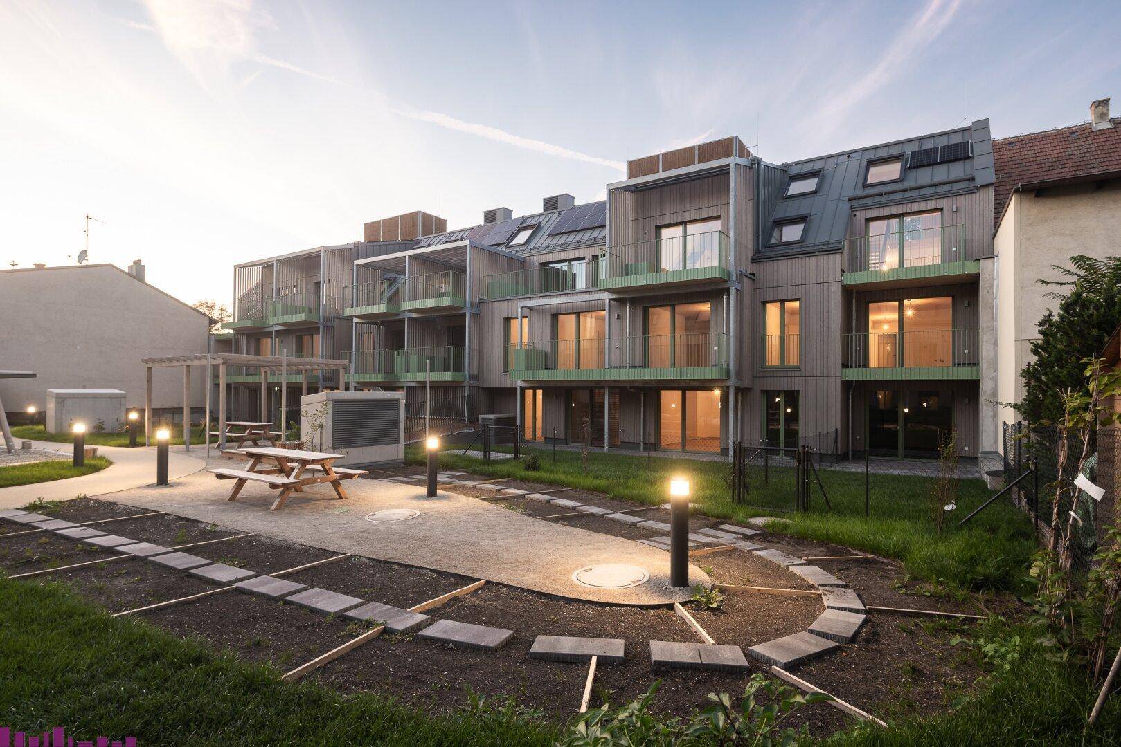Moderne Wohnanlage mit Balkonen, Solarpaneelen und einem beleuchteten Gemeinschaftsgarten mit Sitzbereich.