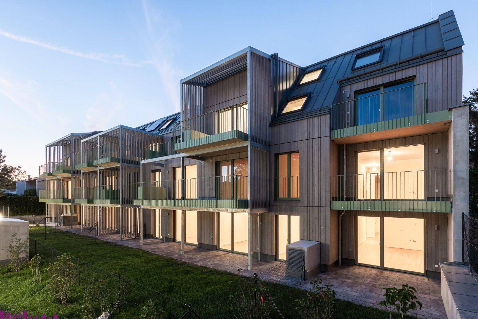 Moderne Wohngebäude bei Dämmerung mit beleuchteten Innenräumen, Balkonen und Terrassenzugang.