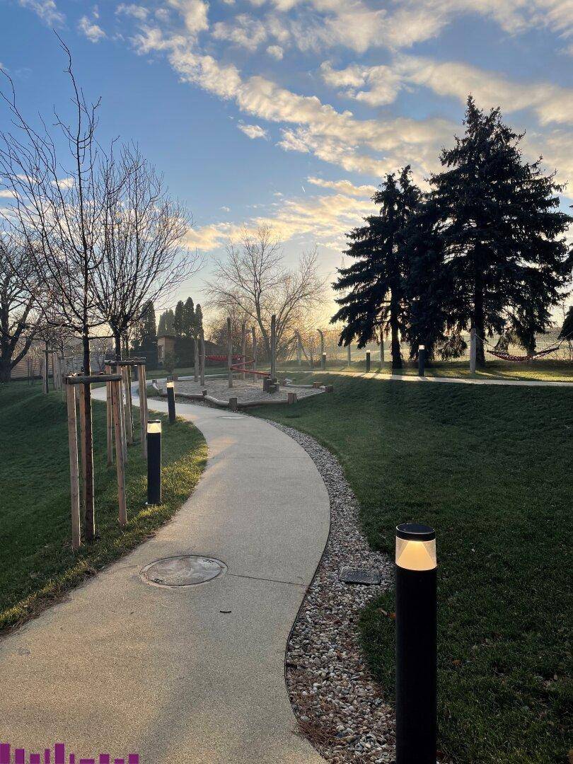 Geschwungener Weg durch eine grüne Parkanlage mit Spielplatz und stimmungsvoller Beleuchtung bei Sonnenaufgang.