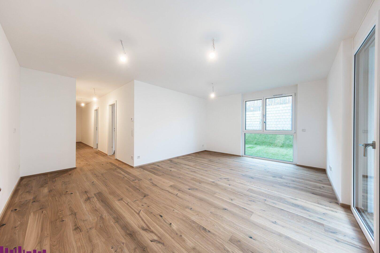 Helles, leeres Wohnzimmer mit breiten Holzplankenböden und Zugang zu einem Gartenbereich.