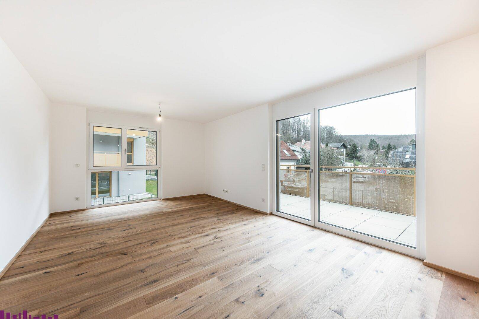 Geräumiges, helles Zimmer mit Holzboden, großen Fenstern und Zugang zu einer Terrasse mit Blick ins Grüne.
