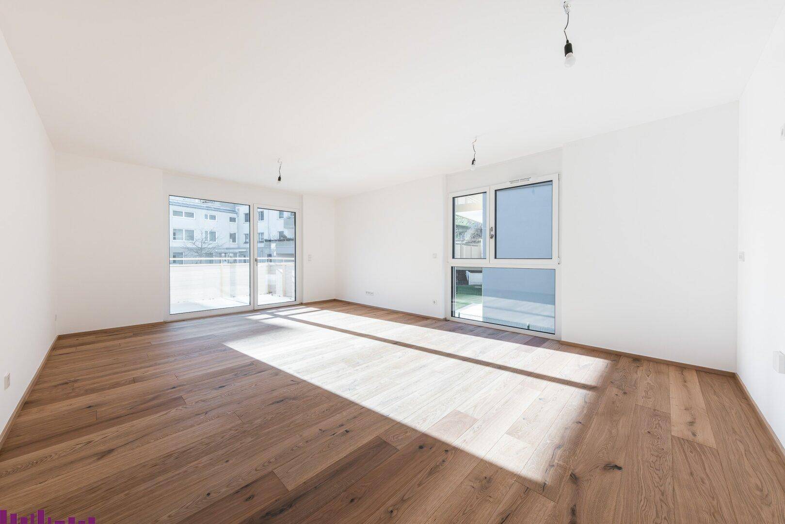 Heller, leerer Wohnraum mit Holzboden, großen Fenstern und Zugang zum Balkon.