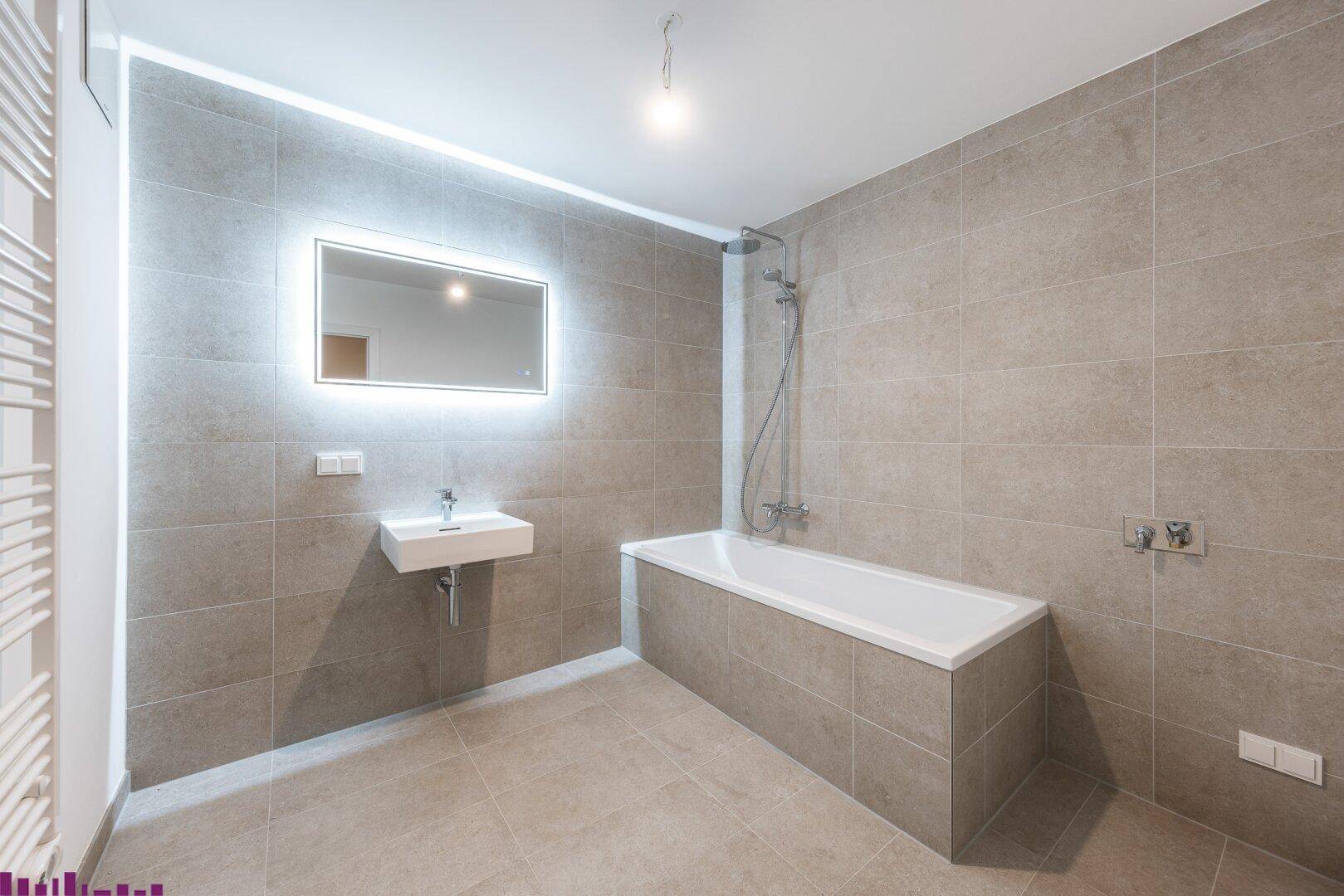 Modernes Badezimmer mit Badewanne, beleuchtetem Spiegel und grauen Großformatfliesen an Wänden und Boden.