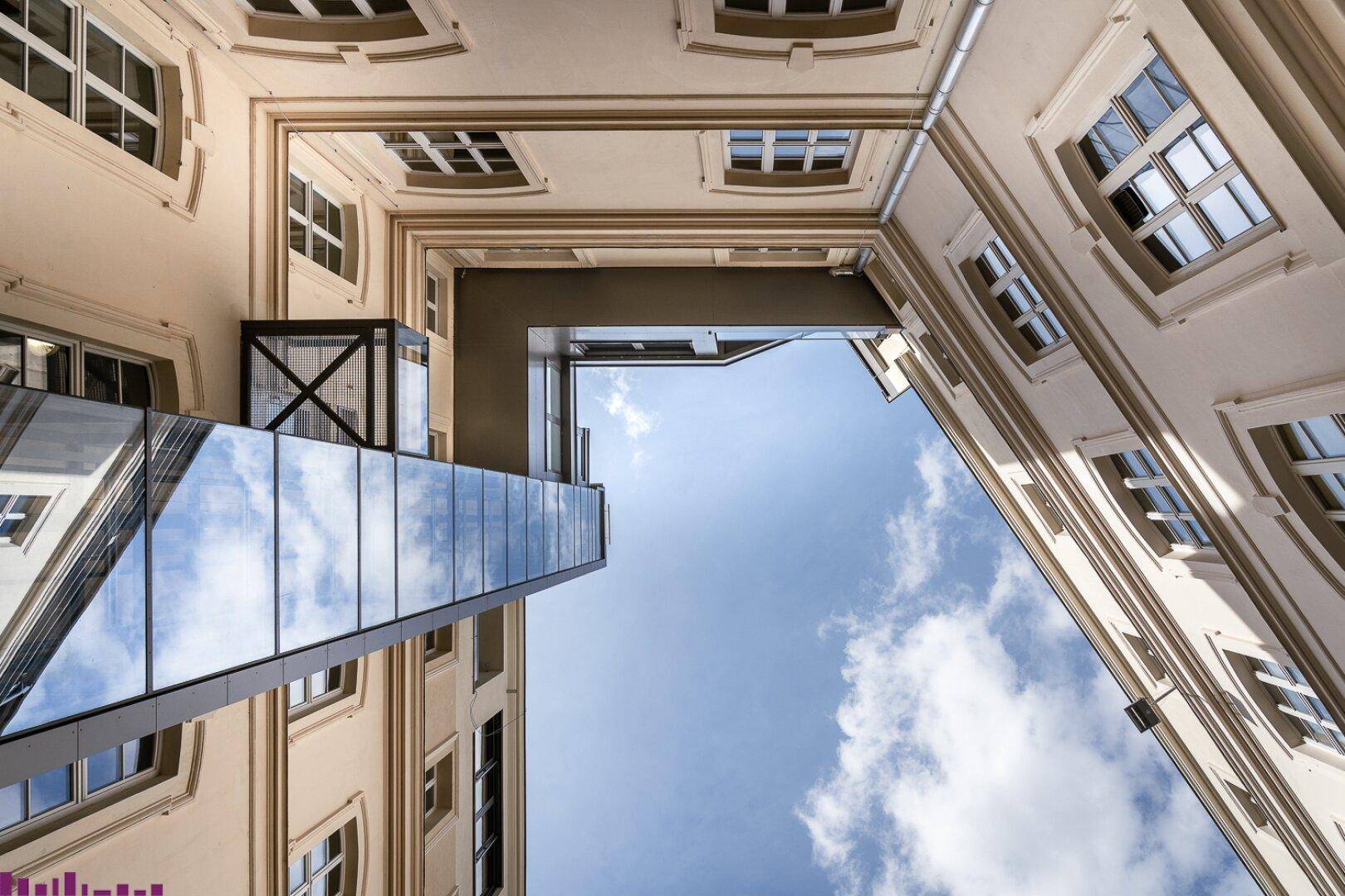 Blick in einen Innenhof mit traditioneller Architektur und einer modernen Glasbrückenkonstruktion unter blauem Himmel.