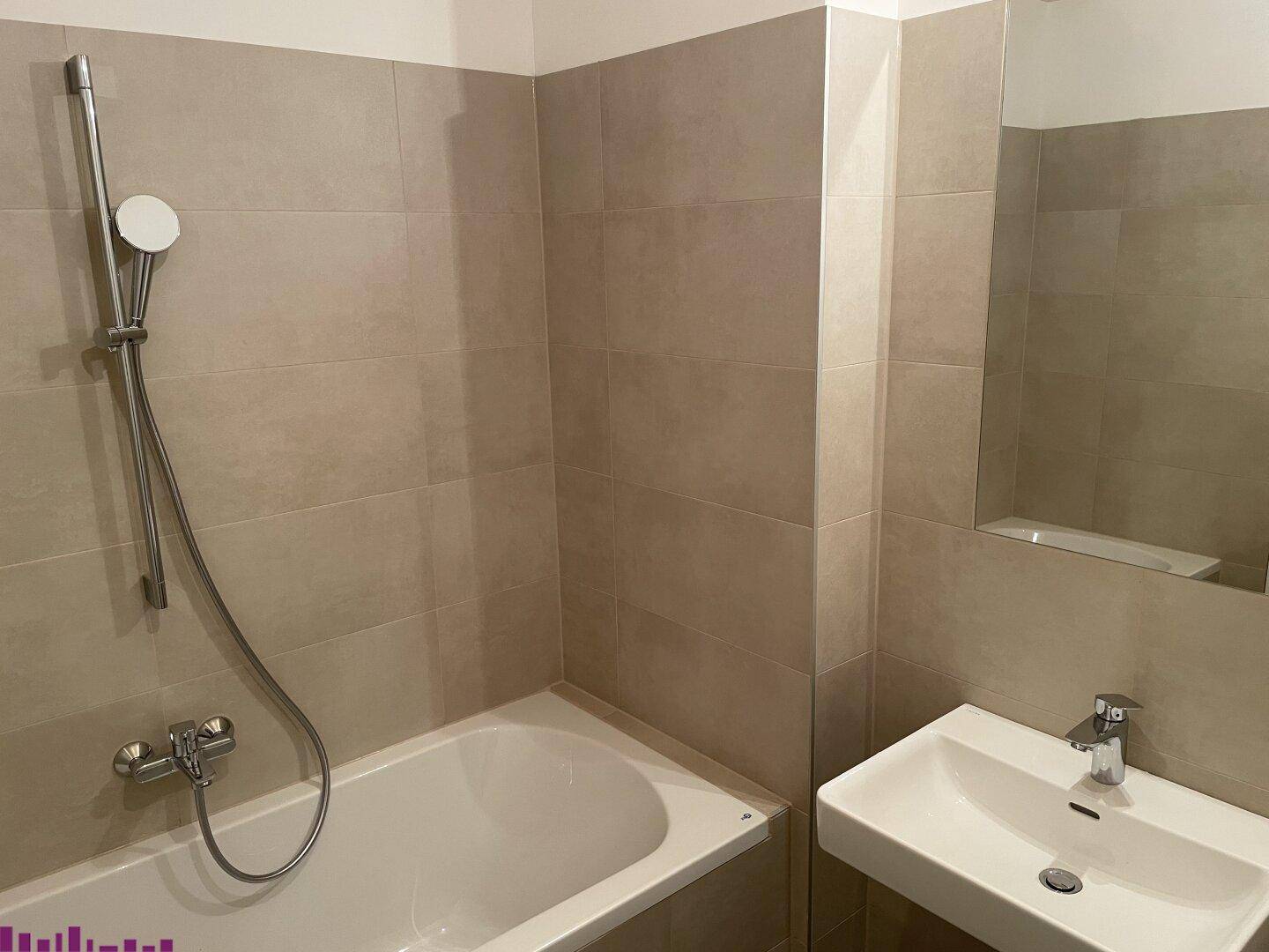 Modernes Badezimmer mit beigefarbenen Fliesen, einer Badewanne mit Duschfunktion und einem Waschbecken mit Spiegel.