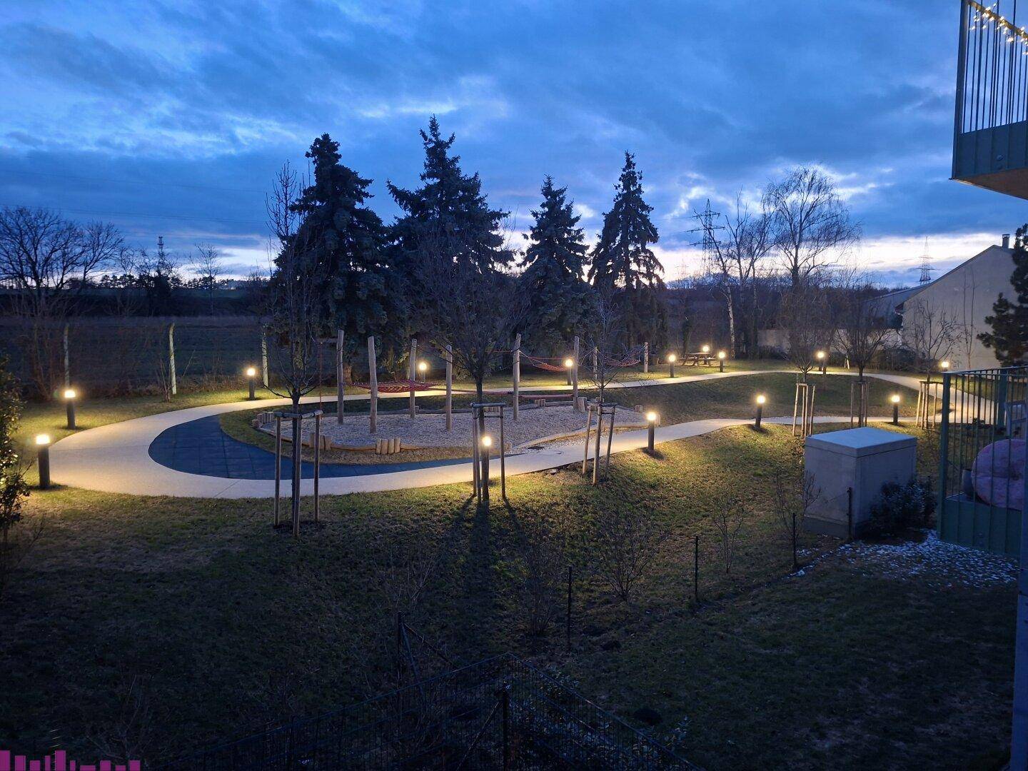 Abendliche Ansicht eines gepflegten Gemeinschaftsgartens mit Spielplatz und beleuchteten Wegen.