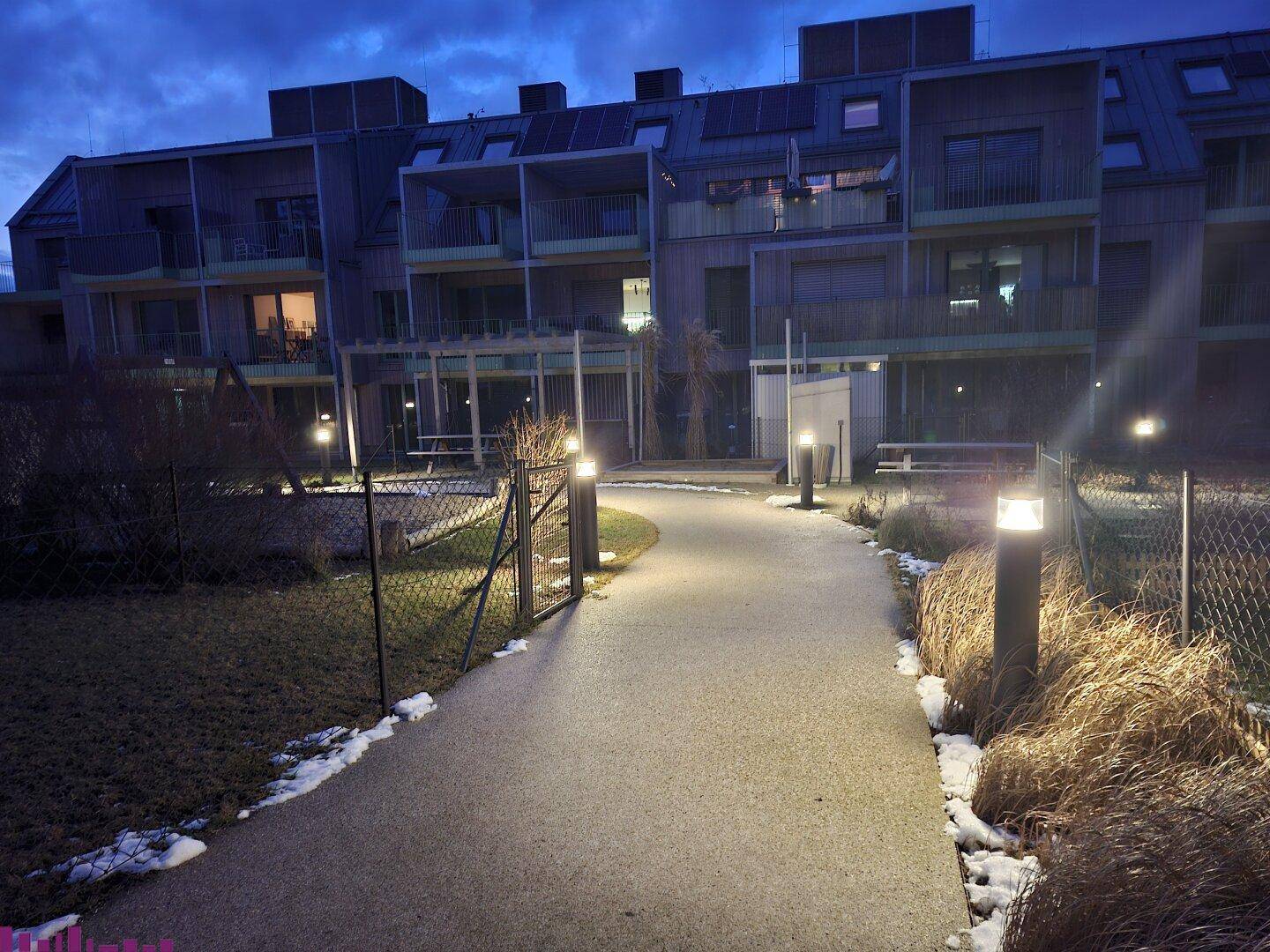 Moderne Wohnanlage bei Dämmerung mit Balkonen, Photovoltaikanlage und beleuchtetem Zugangsweg.