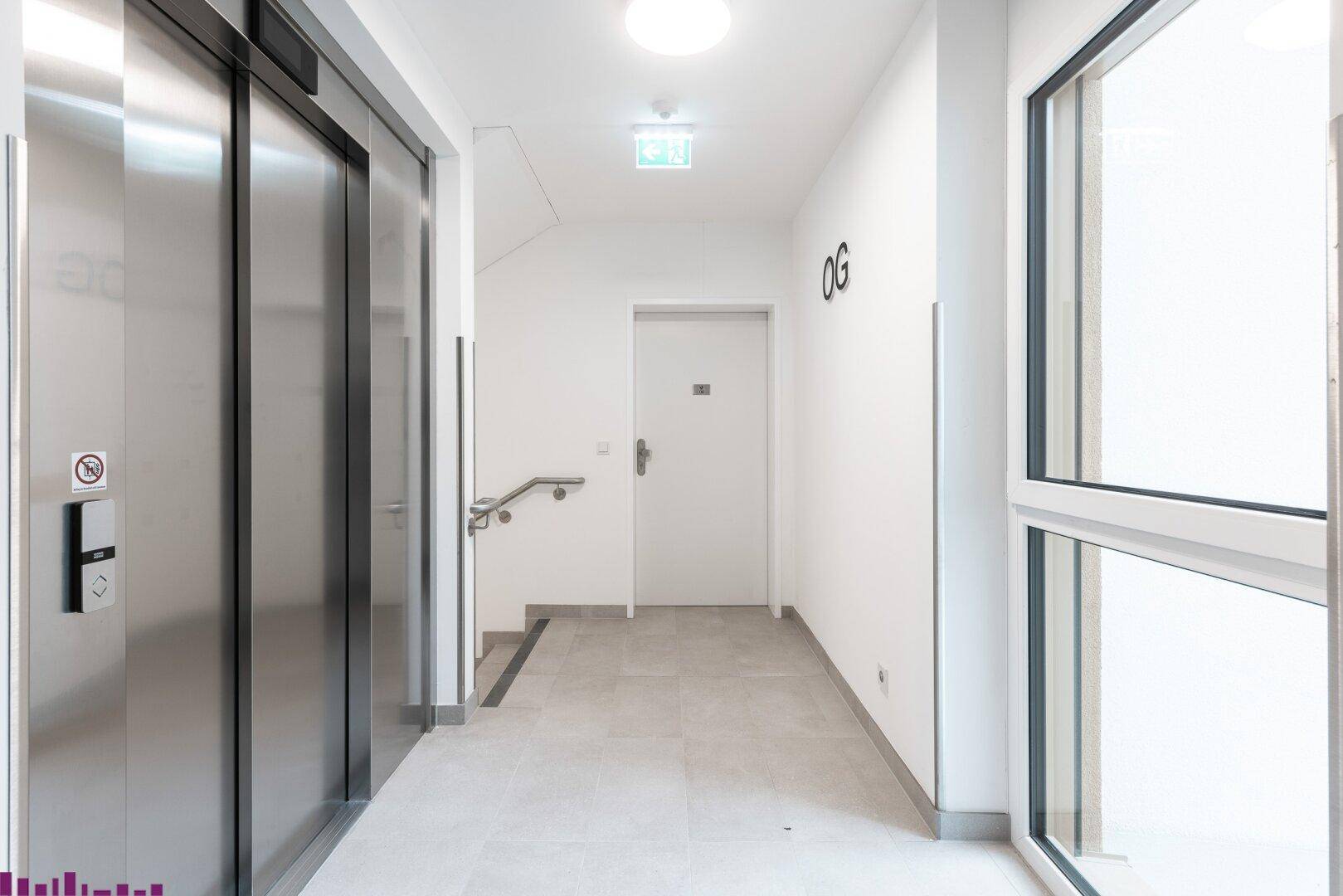 Heller Flur mit Aufzug, Treppenhaus und Fenster im Obergeschoss.