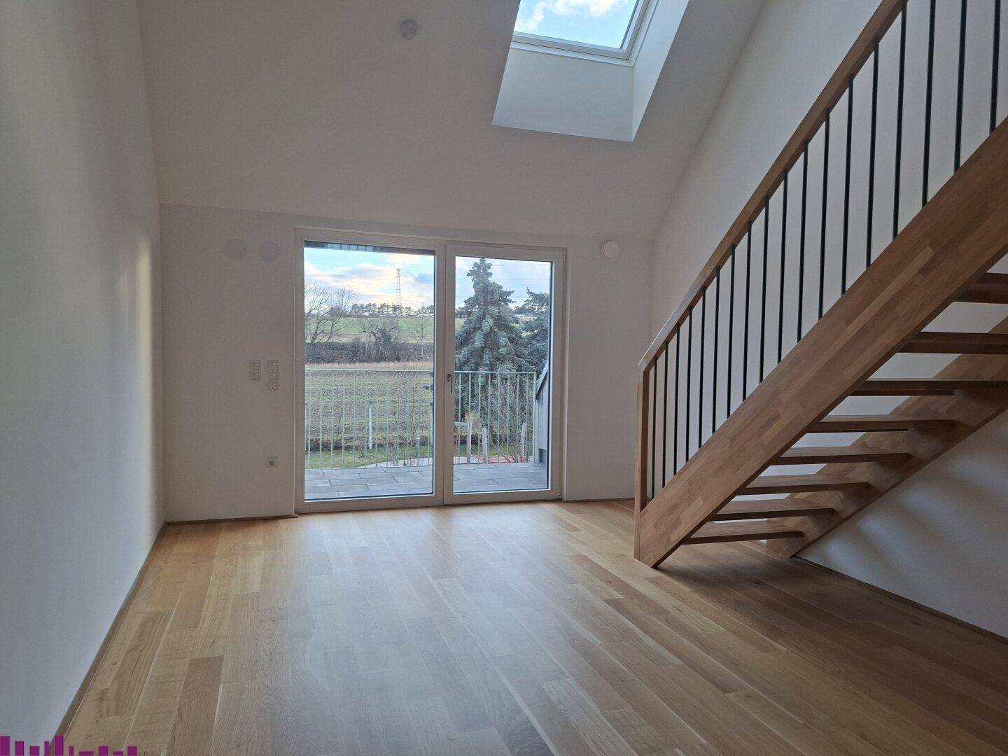 Leerer, heller Raum mit Holzparkett, offener Holztreppe und großem Fenster mit Zugang nach draußen.