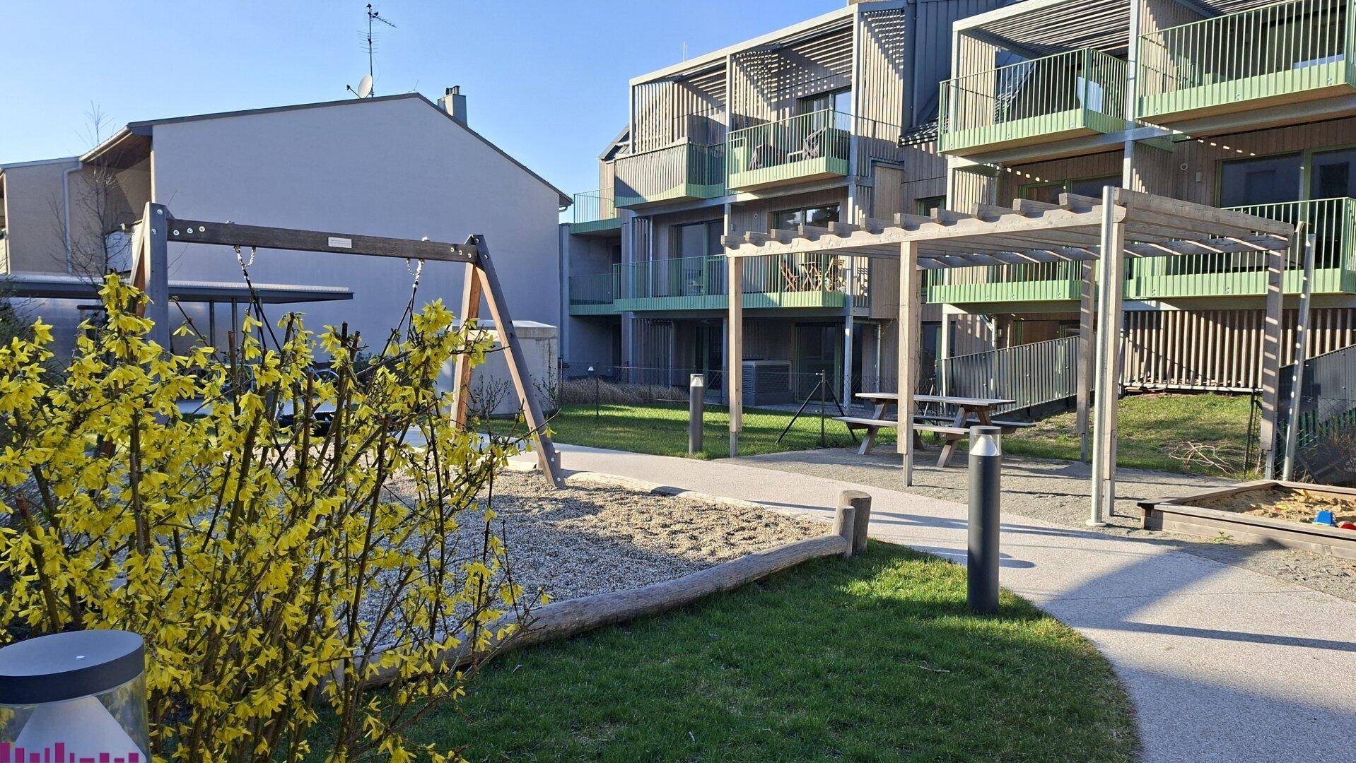 Gemeinschaftsgarten mit Spielplatz, Schaukeln und Pergola vor modernen Wohngebäuden.