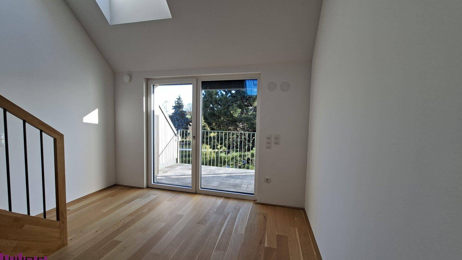 Heller Raum mit Holzboden, Treppenzugang und großer Balkontür mit Blick ins Grüne.