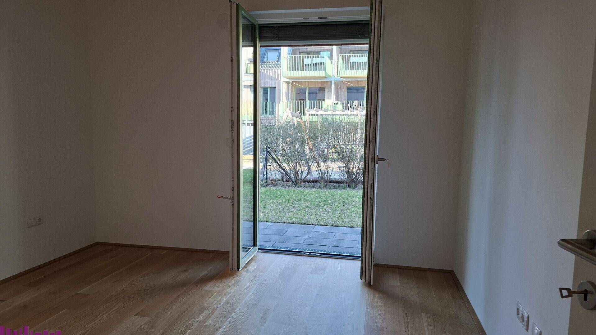 Heller, leerer Raum mit Parkettboden und einer Tür, die direkten Zugang zum Garten bietet.