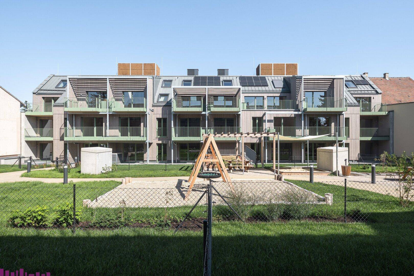 Moderne Wohnanlage mit Balkonen und einem Spielplatz im begrünten Innenhof unter blauem Himmel.