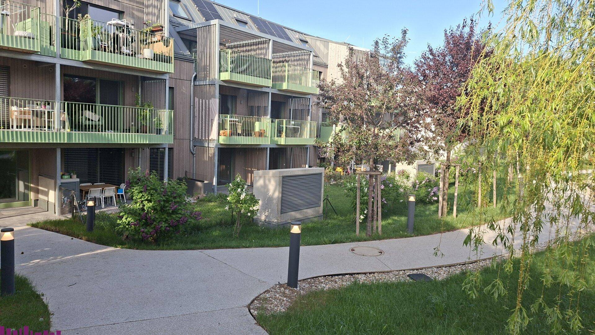 Gepflegte Gartenanlage mit Gehwegen und Beleuchtung vor der modernen Wohnanlage, umgeben von Bäumen.