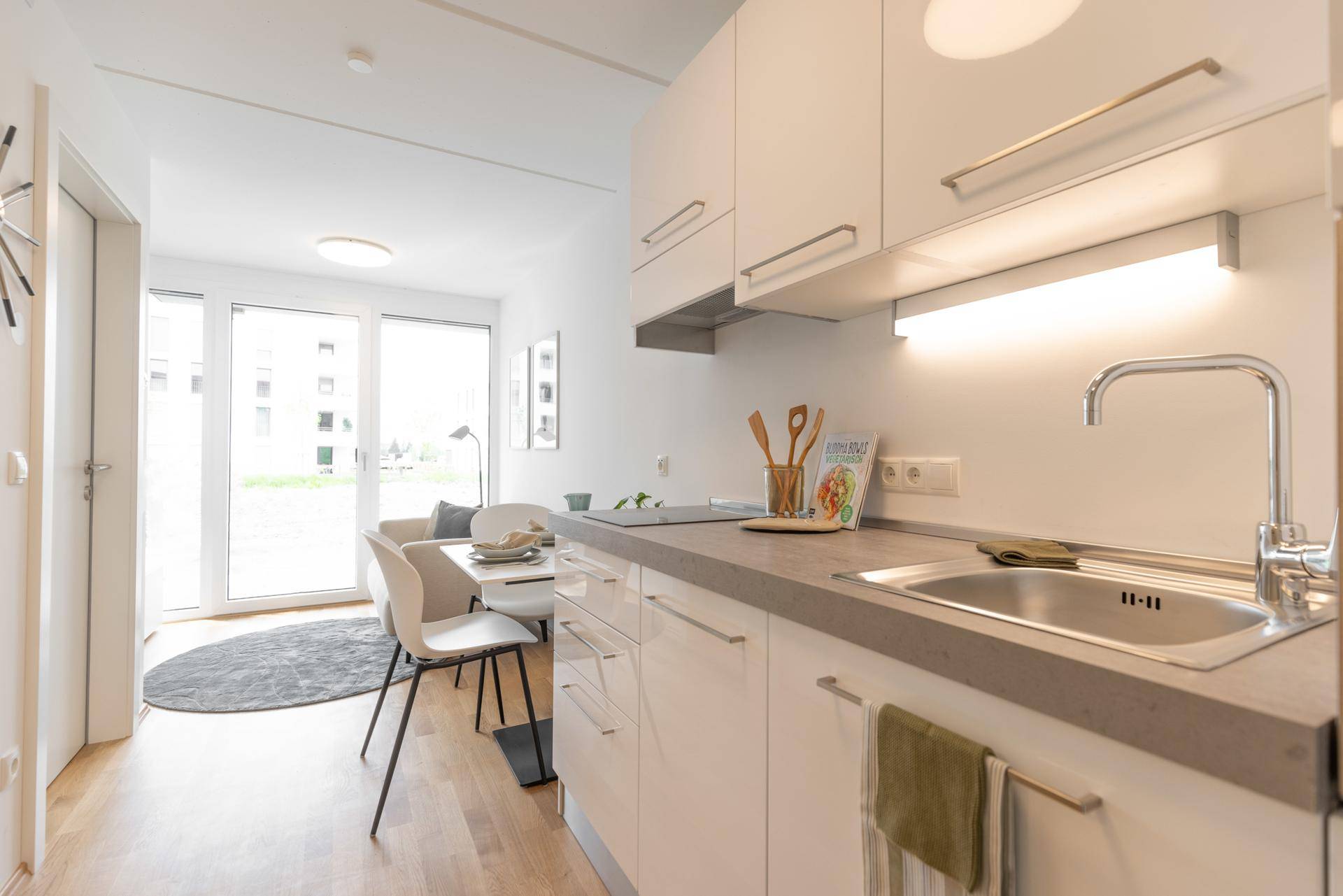 Moderne Küchenzeile mit Spüle, Kochfeld und weißen Schränken, offen zum Essbereich und Wohnzimmer.