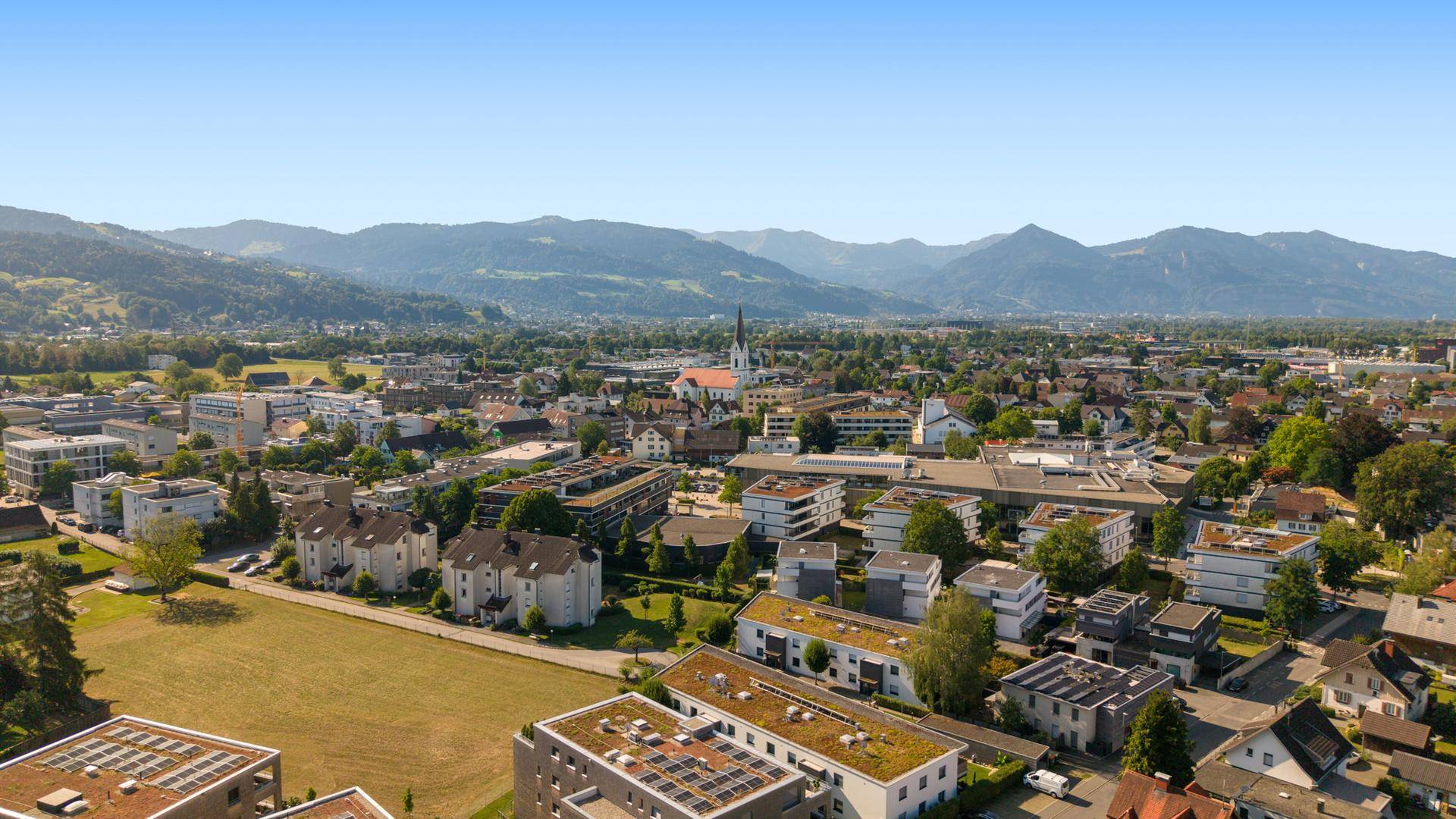 Luftaufnahme einer Stadt mit Wohngebäuden, Grünflächen und Bergen im Hintergrund.