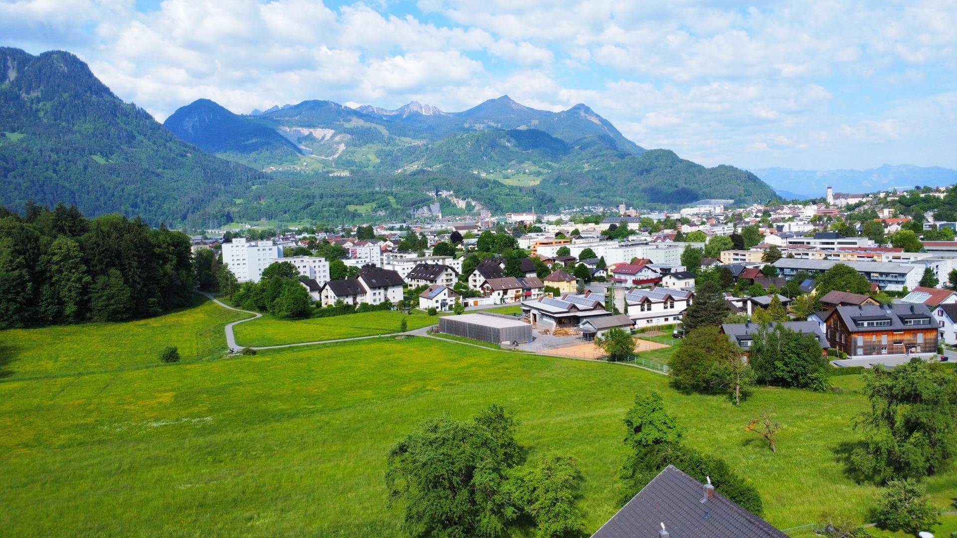 Weitläufige Landschaftsaufnahme mit grünen Feldern, einer Stadt und majestätischen Bergen im Hintergrund.