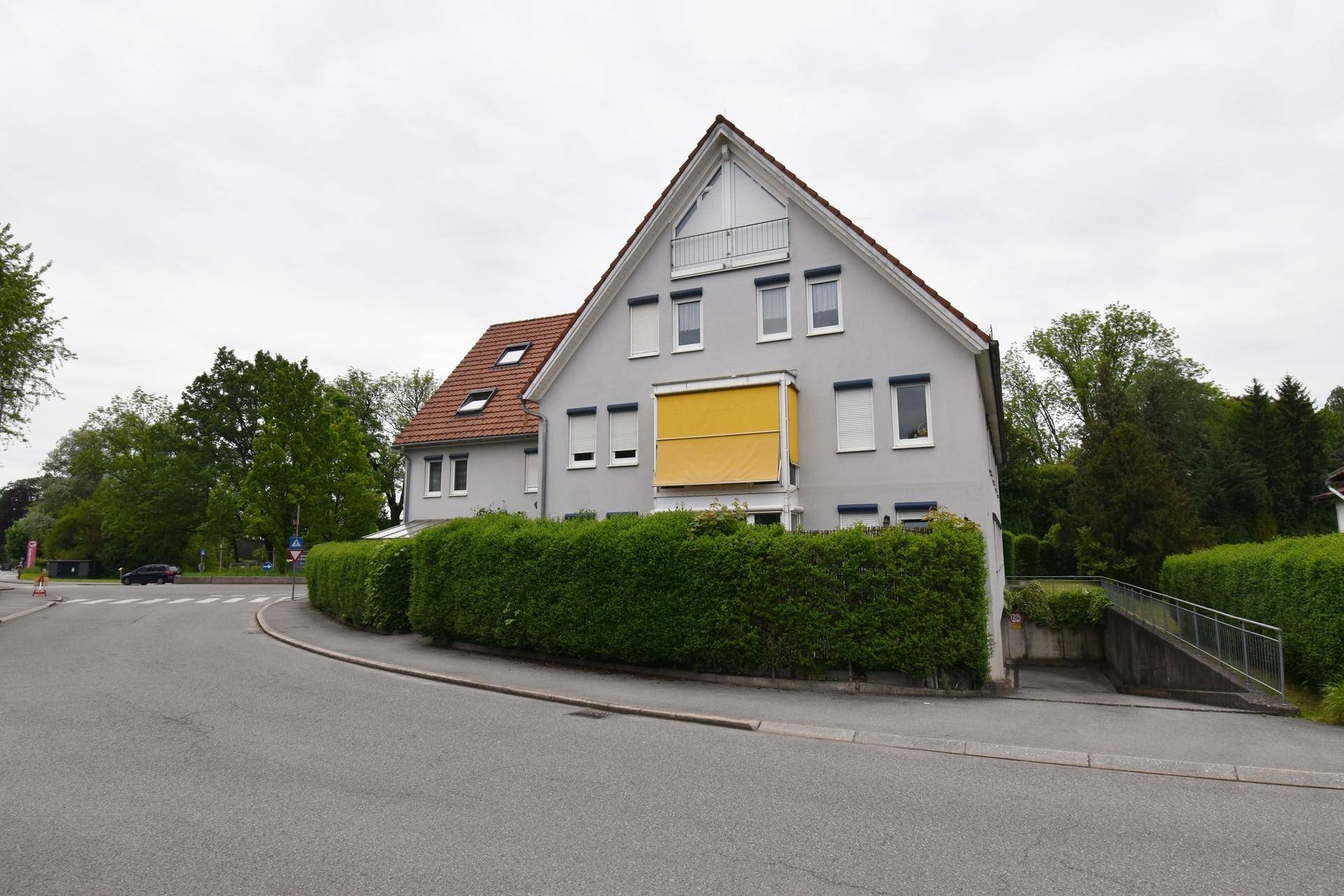 Mehrfamilienhaus mit grauer Fassade, gelbem Sonnenschutz und gepflegter Hecke.