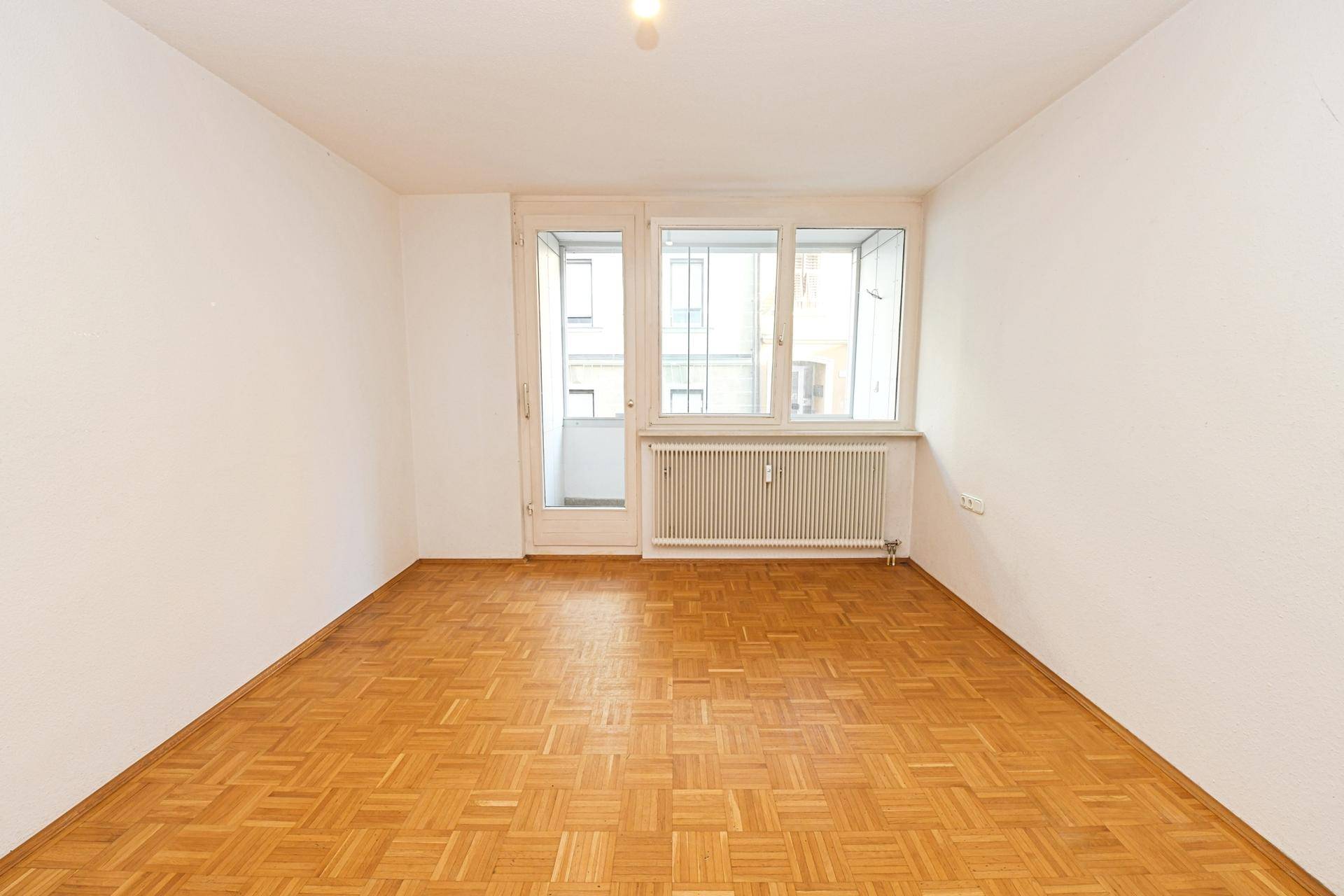 Helles Zimmer mit Fischgrätparkettboden und Zugang zu einem kleinen Balkon.