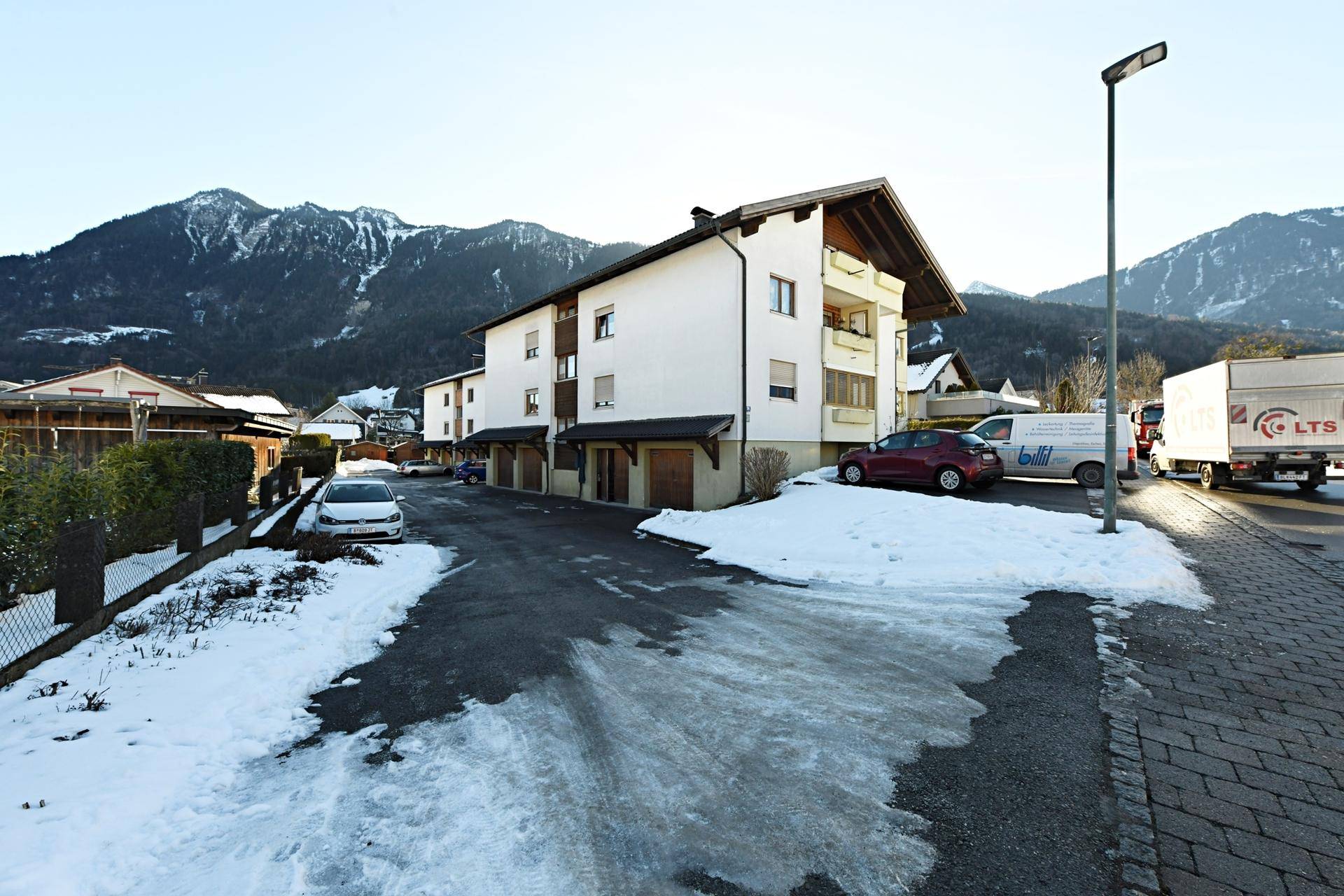 Mehrfamilienhaus in ländlicher Umgebung mit verschneiter Zufahrt und Bergblick.