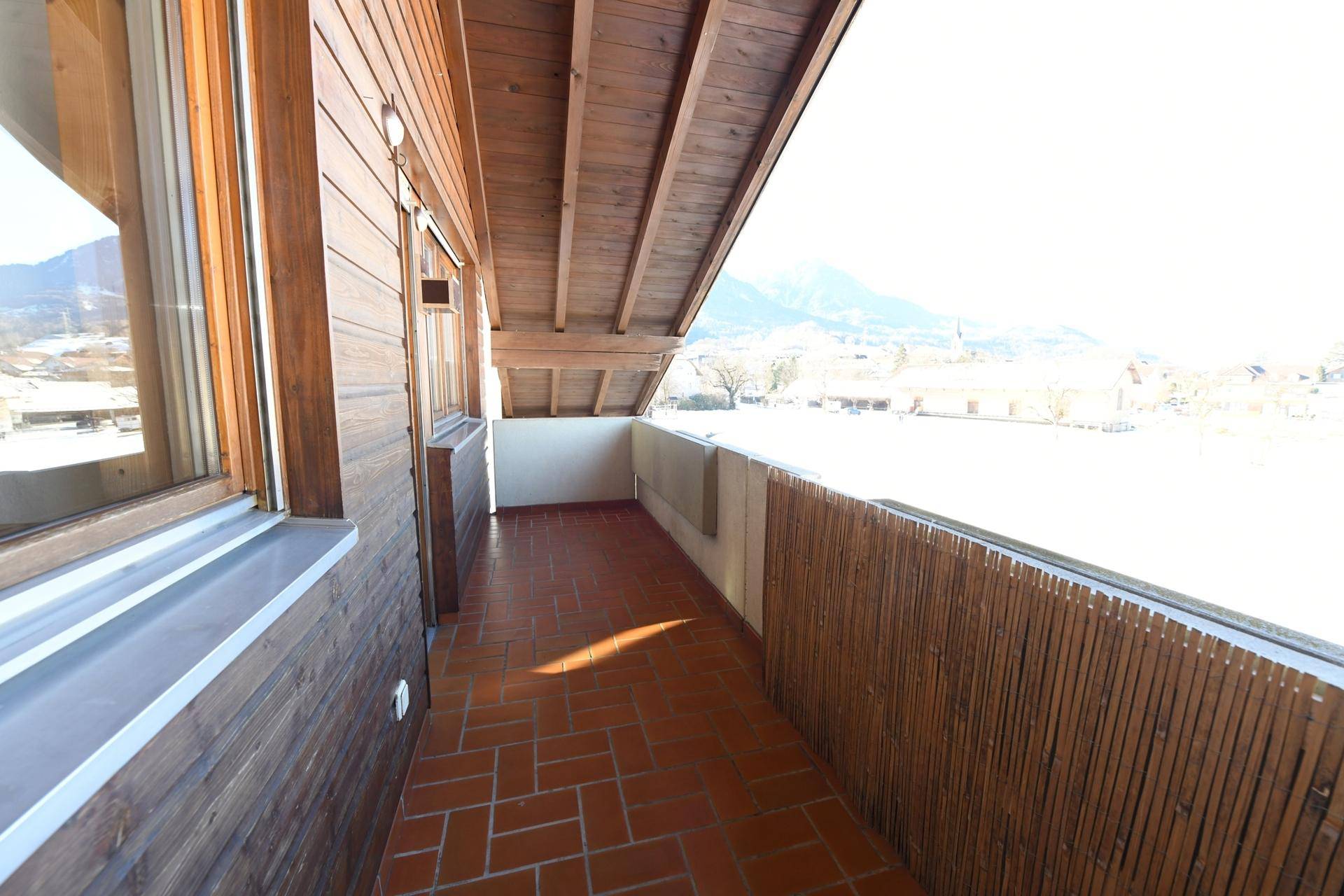 Sonniger Balkon mit Terrakotta-Fliesen und Holzverkleidung, umgeben von Bergen.