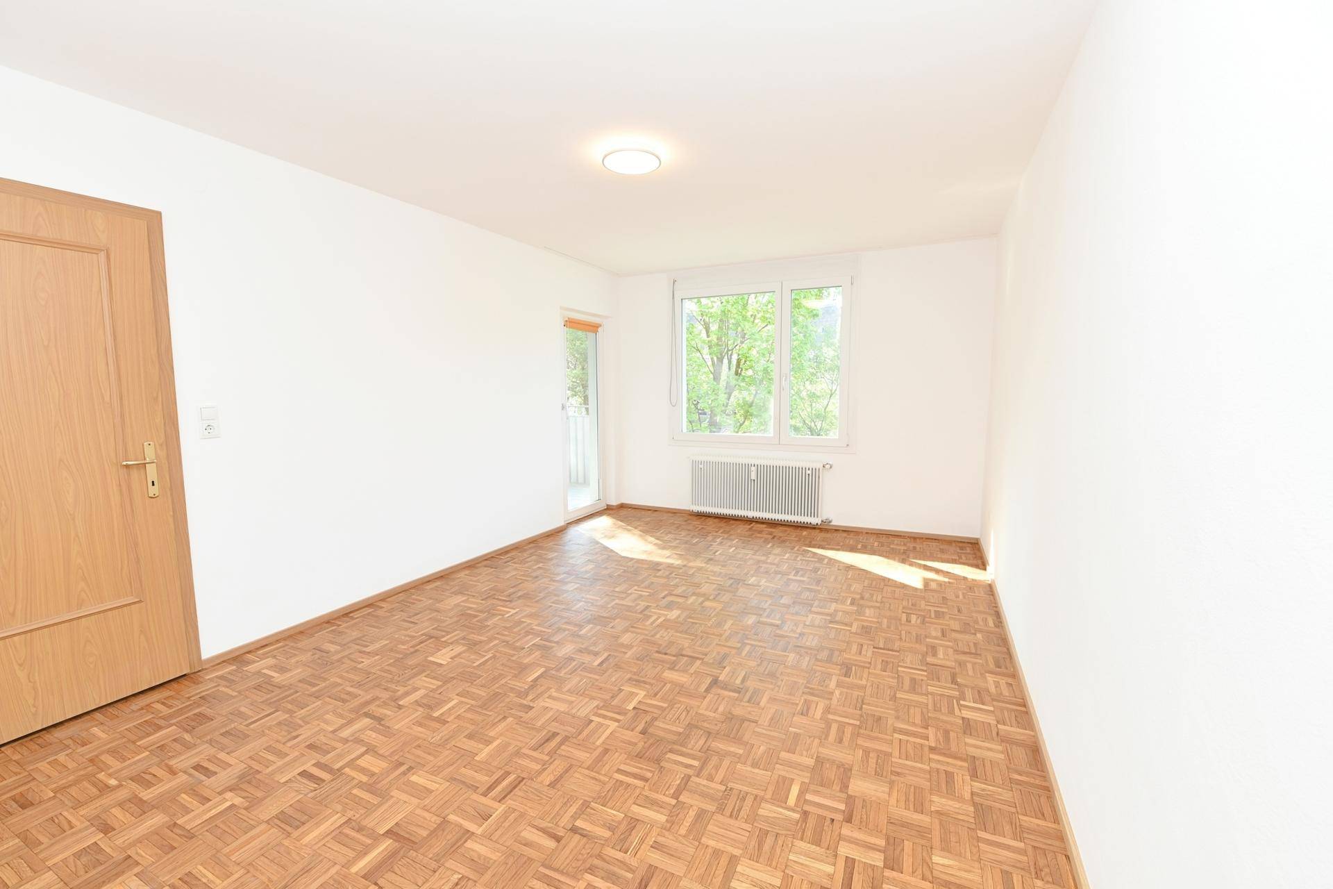 Helles Zimmer mit Parkettboden und Balkonzugang, bietet viel Tageslicht und frische Luft.