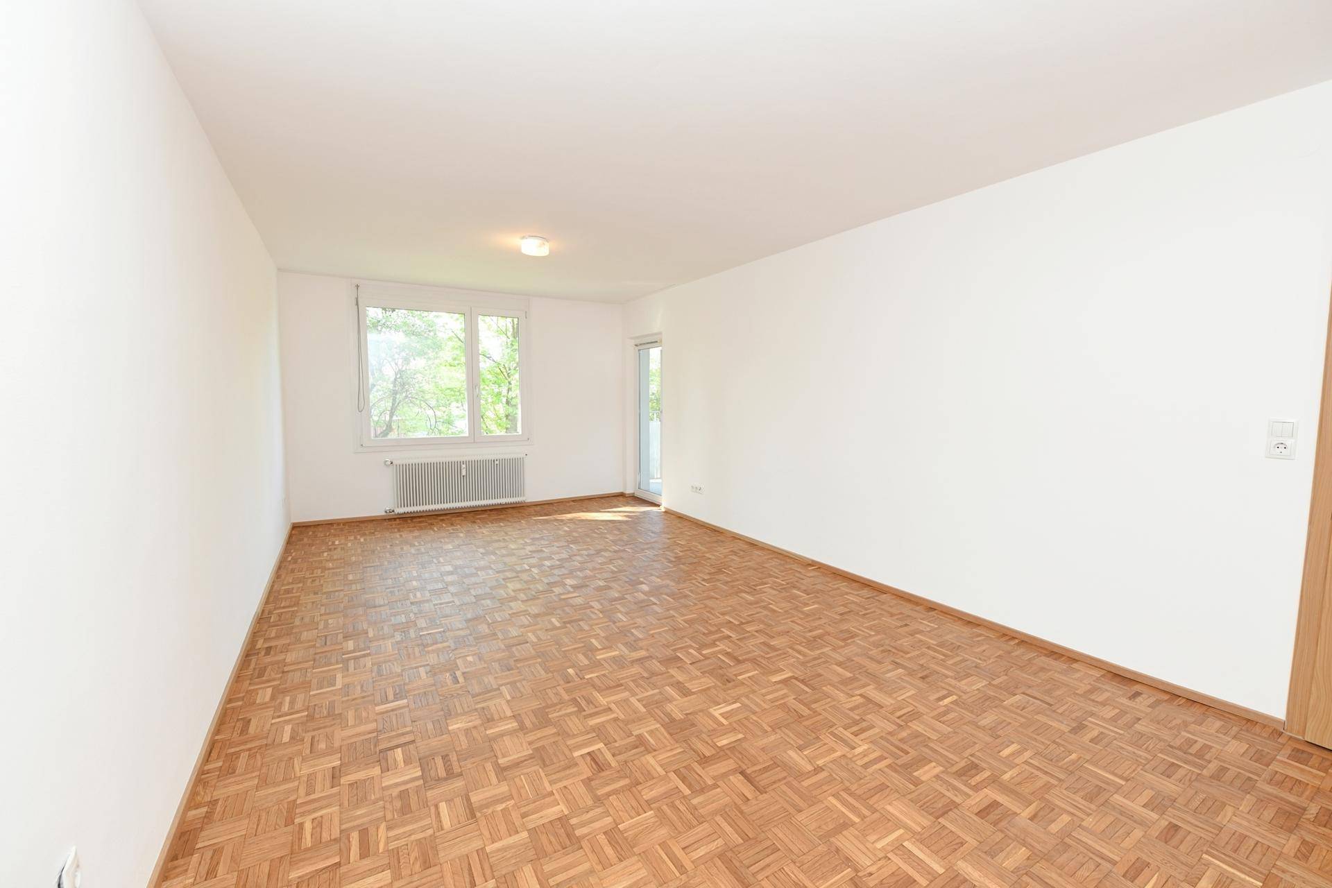 Geräumiges, leeres Zimmer mit Parkettboden und Zugang zu einem Balkon, hell und einladend.