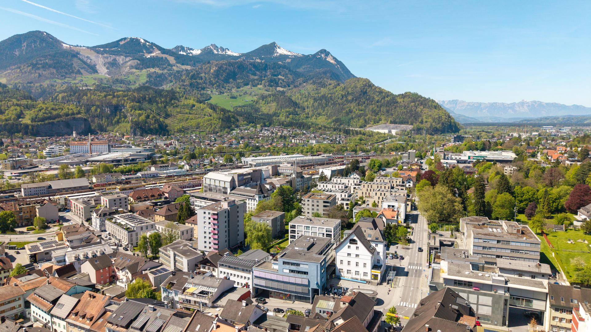 Panoramablick aus der Luft auf eine Stadt mit grünen Hügeln und Bergen am Horizont.