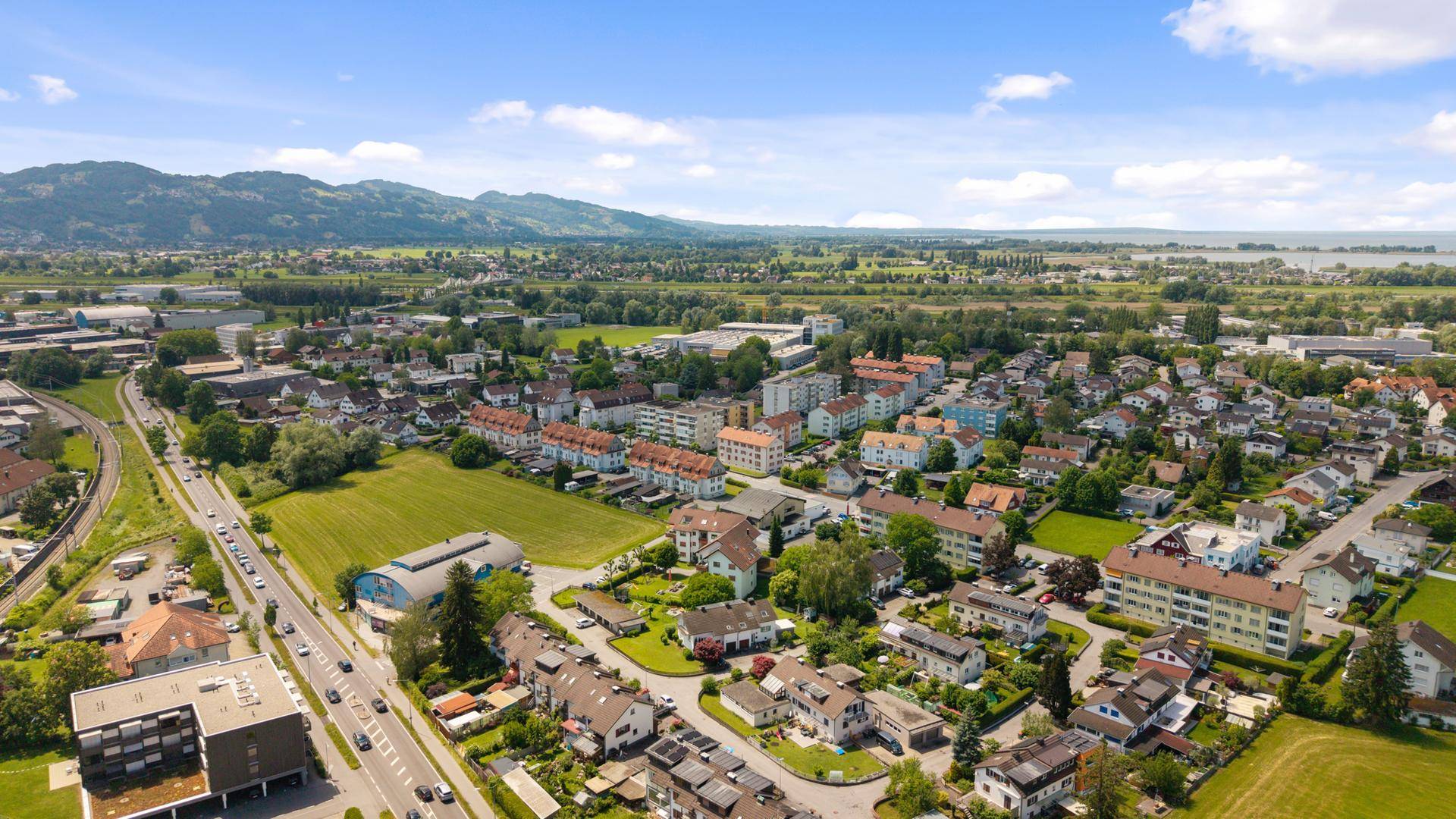 Weitläufige Luftaufnahme einer Siedlung mit grünen Feldern und Bergen im Hintergrund unter blauem Himmel.