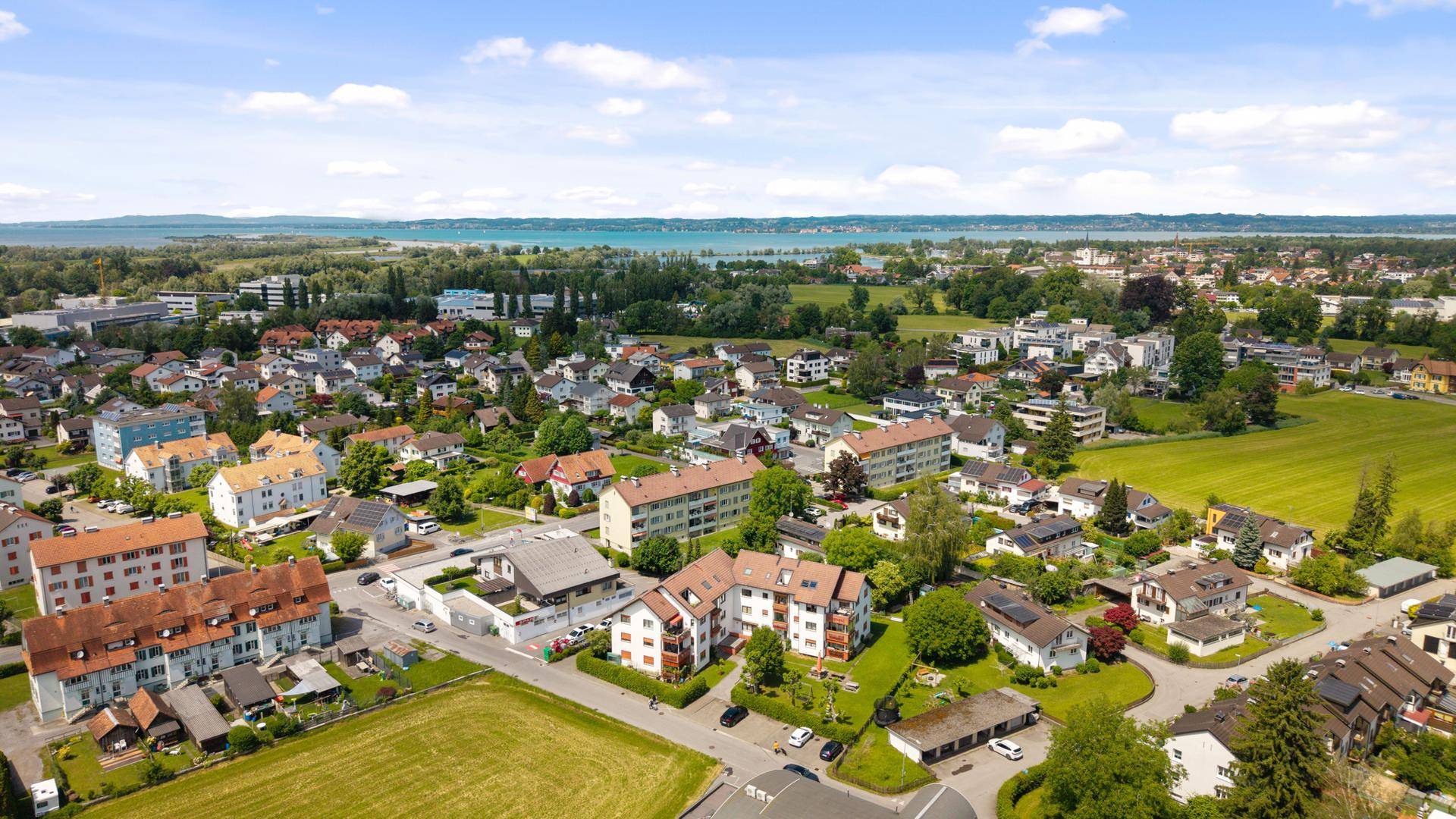 Panoramische Luftaufnahme einer Stadt mit Wohngebäuden, Feldern und einem See am Horizont.
