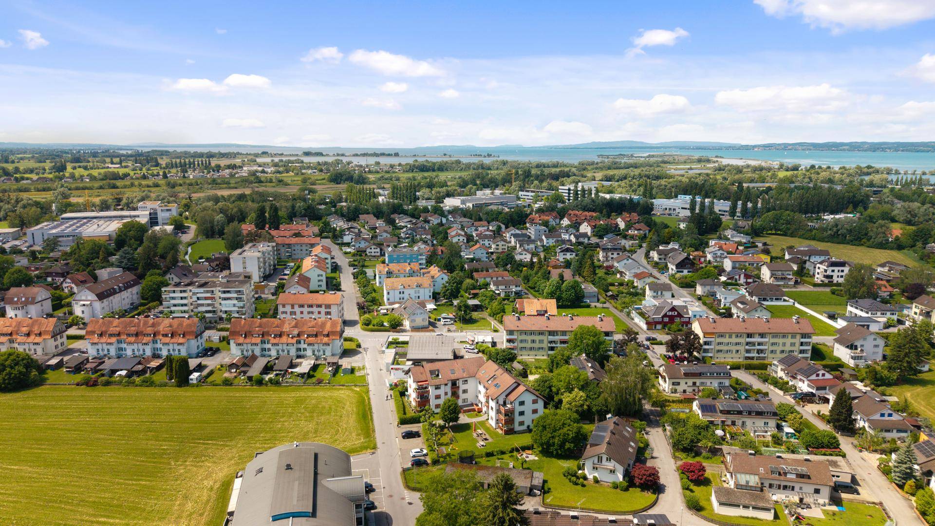 Weitläufige Luftaufnahme einer Wohnsiedlung mit Feldern und einem See im Hintergrund.