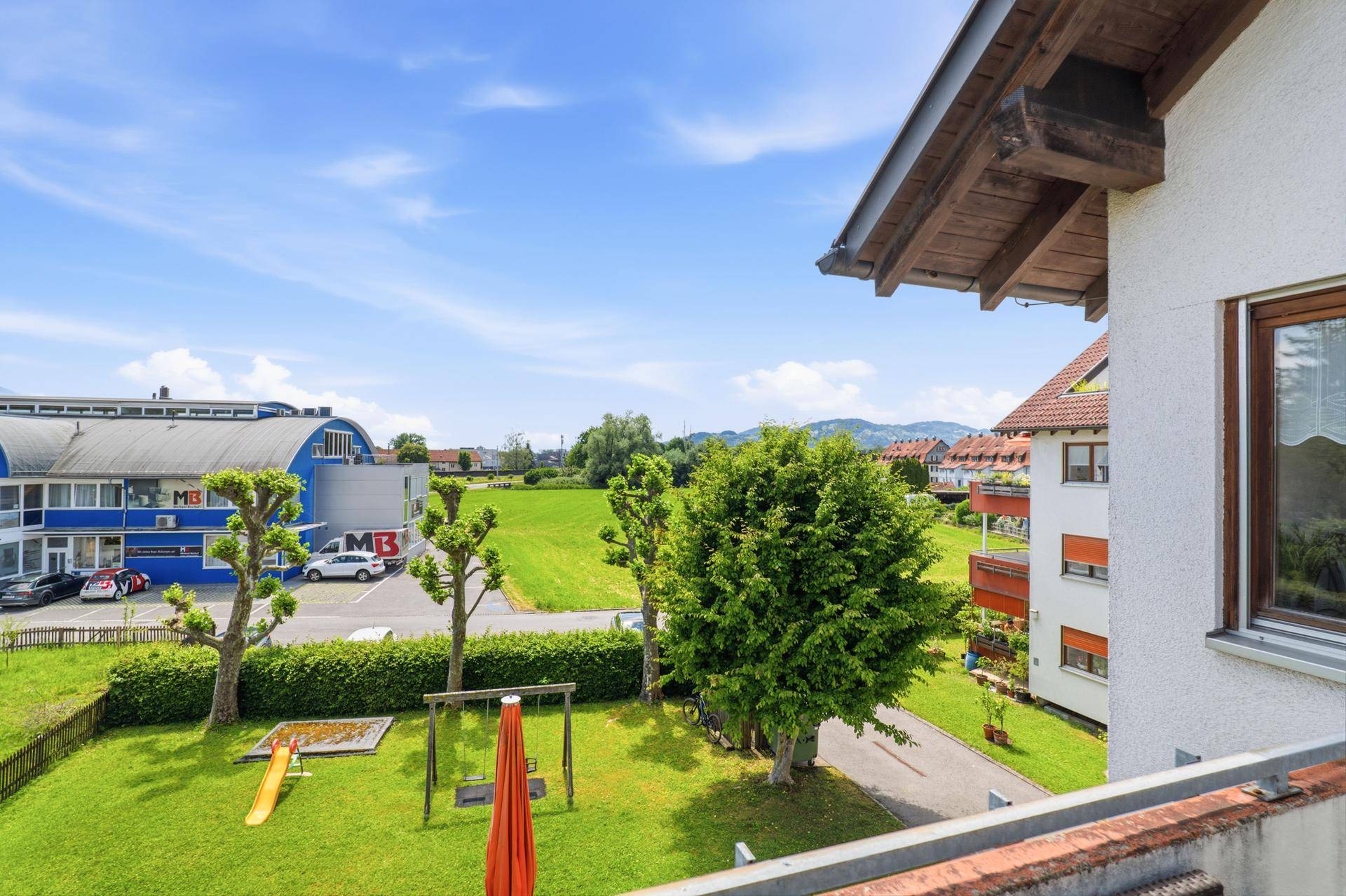 Blick vom Balkon auf einen Gemeinschaftsgarten mit Spielplatz und grüne Felder.