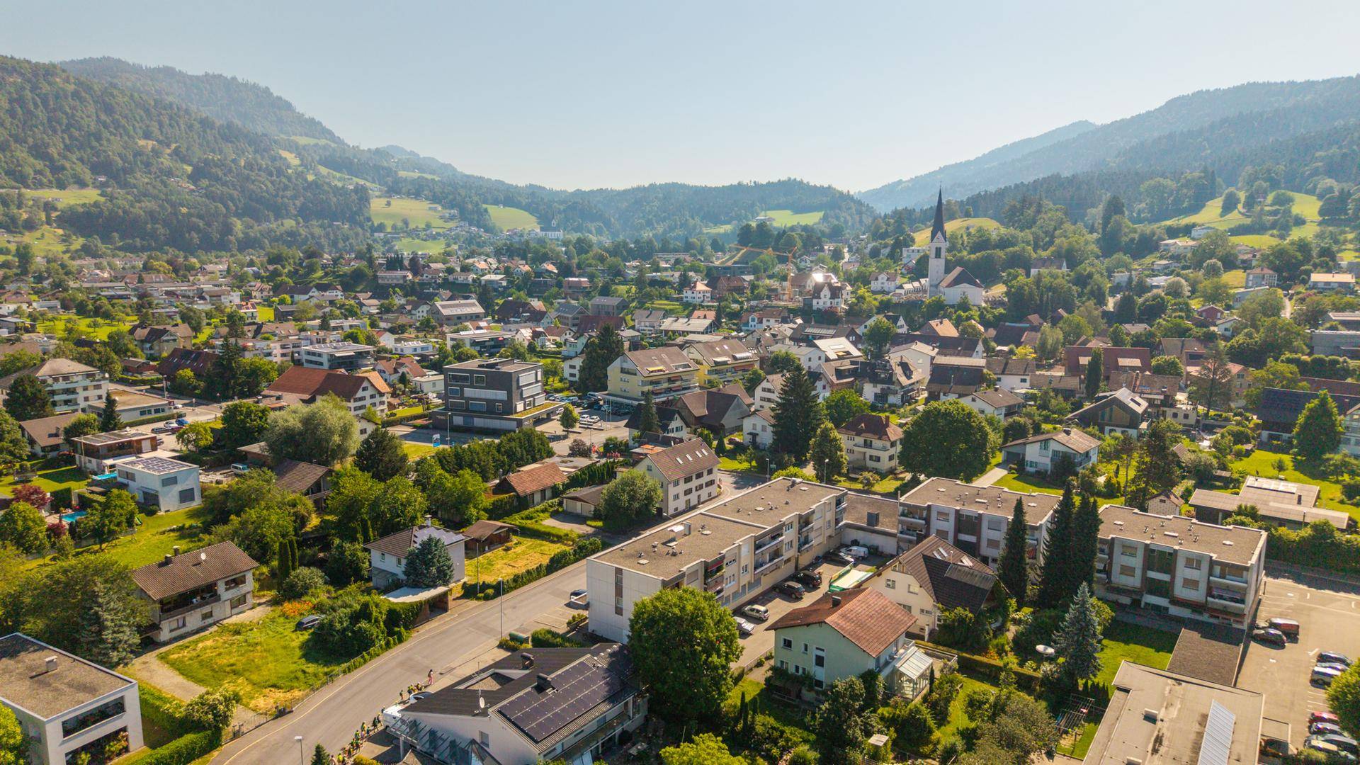 Panoramische Luftaufnahme einer Kleinstadt mit vielen Wohnhäusern und einer Kirche vor bergiger Kulisse.
