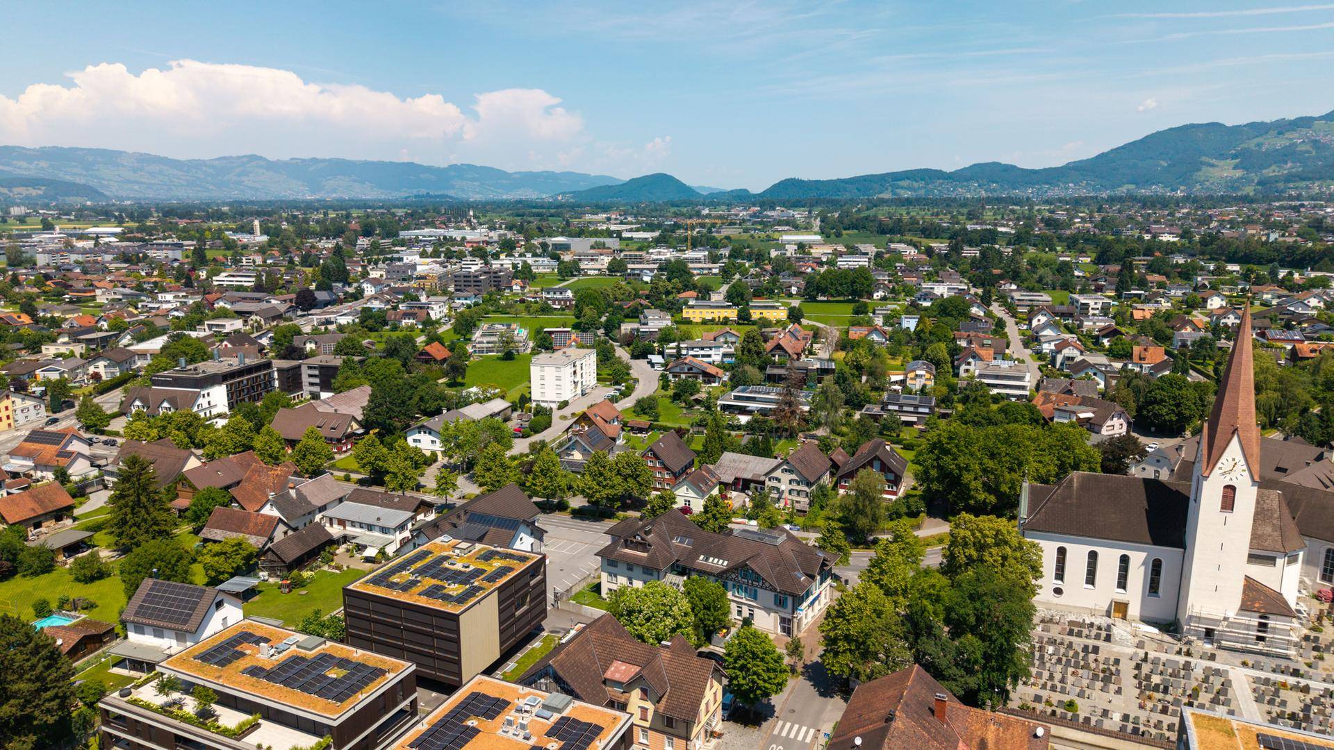 Luftaufnahme einer Stadt mit Wohngebäuden, viel Grün und Bergen im Hintergrund.