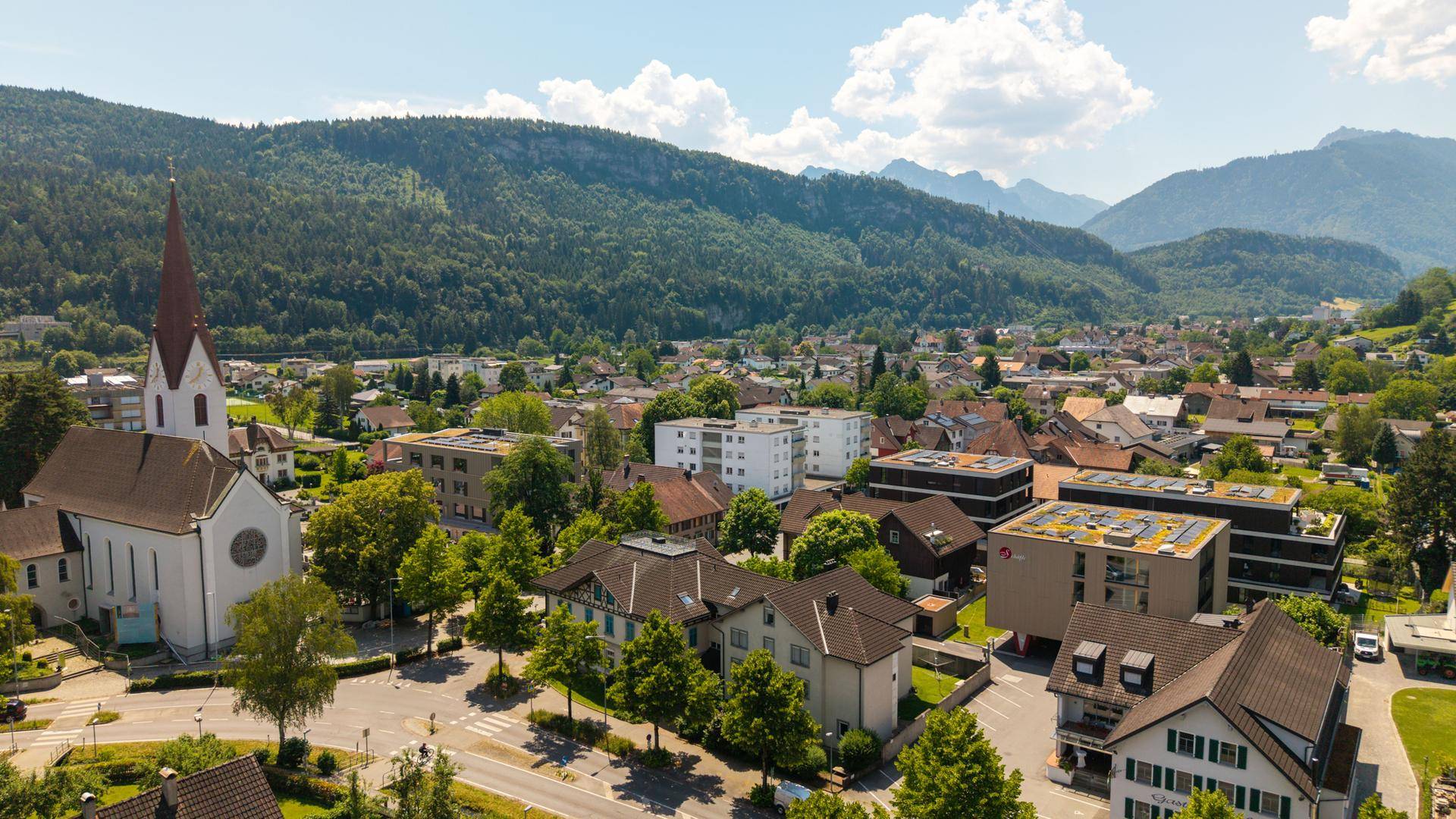 Luftaufnahme einer Wohngegend mit Kirche, umgeben von bewaldeten Hügeln und Bergen.